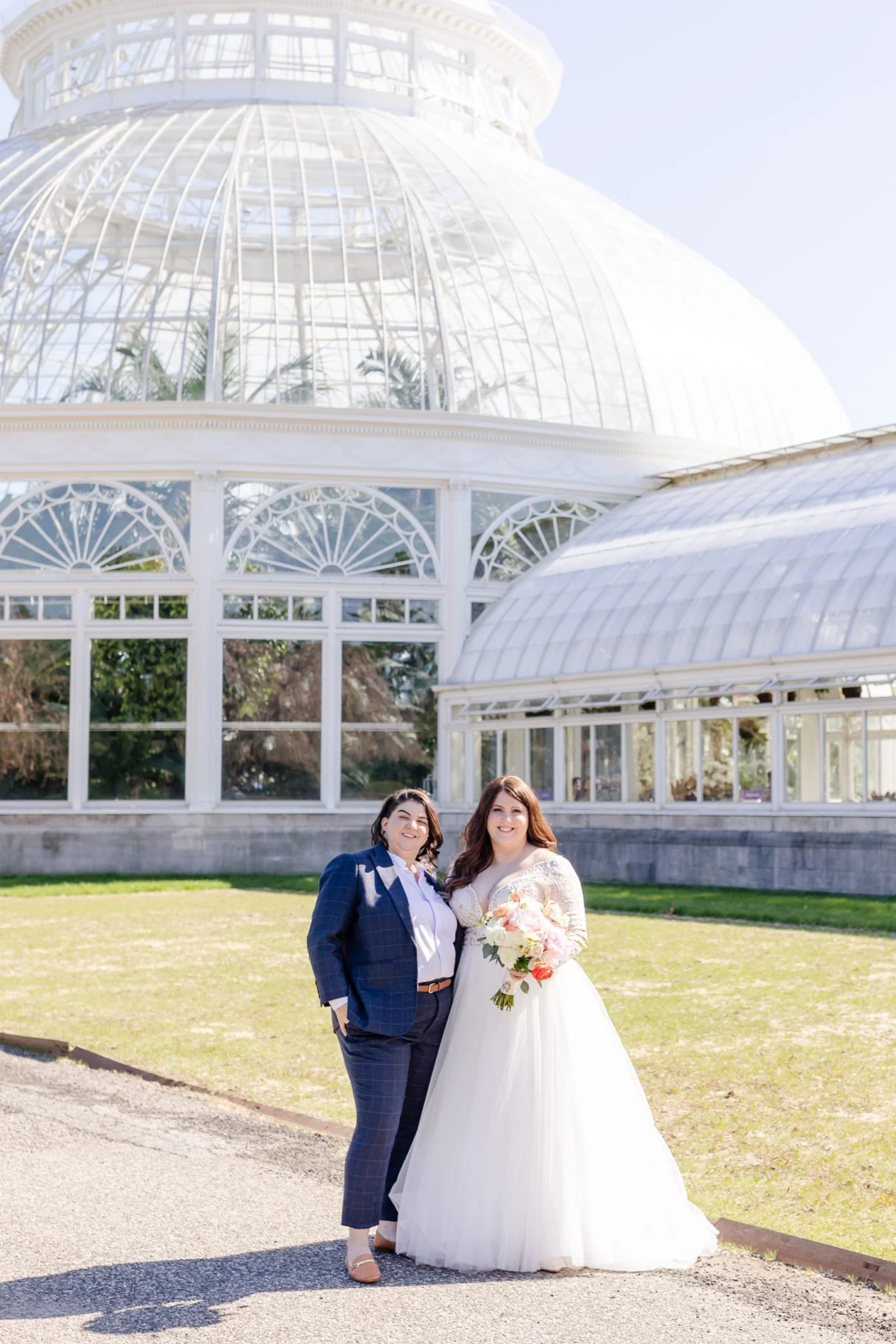 Two women on their wedding day, one in a wedding gown holding a bouquet, and the other in a dark blue checkered suit, standing outdoors in front of a large white greenhouse structure with glass and metal framing, on a sunny day.