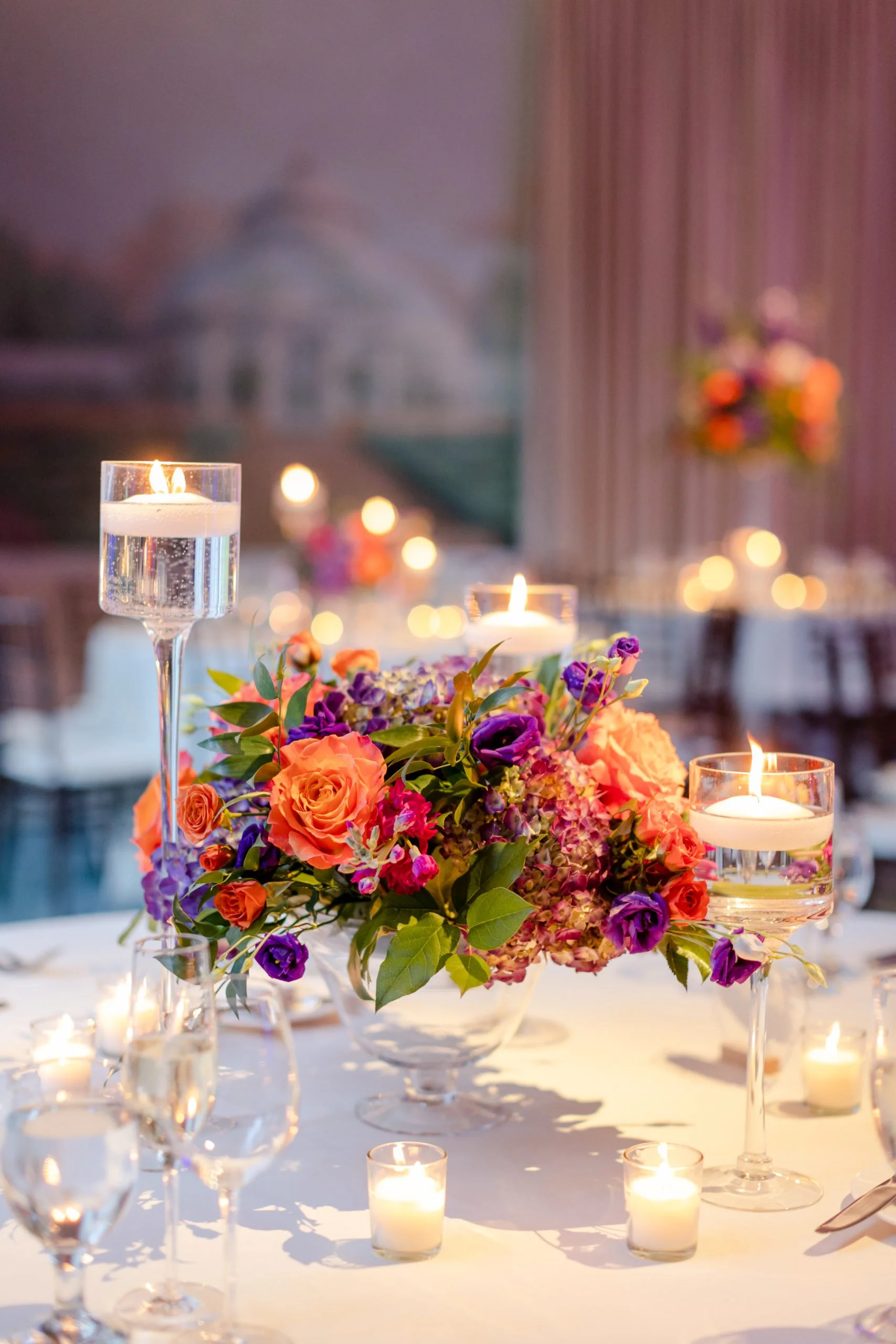 A romantic table setting with a colorful flower centerpiece, lit candles, and elegant glassware, with blurred background of a building and drapes.
