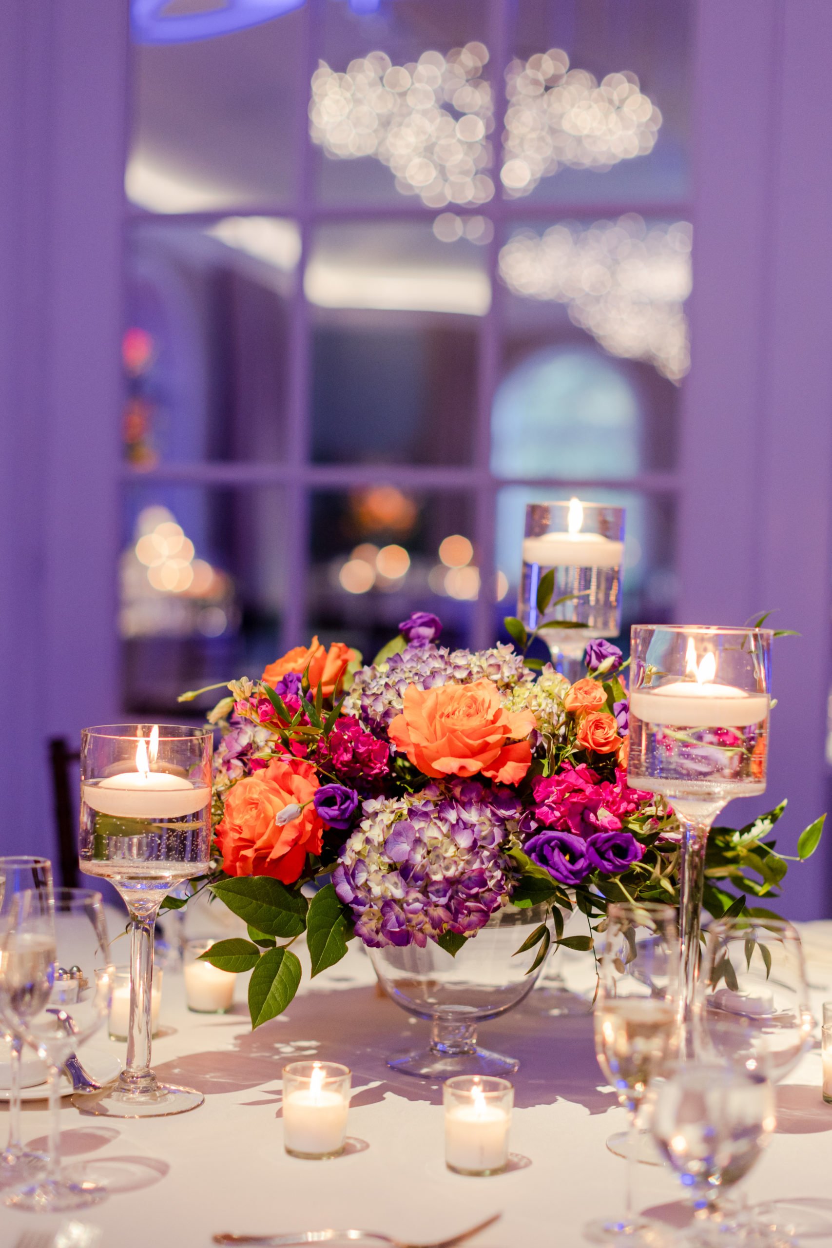 A floral centerpiece with orange, purple, and pink flowers on a table surrounded by tall glass candle holders with lit candles. The background is blurred with purple lighting and decorative bokeh lights.