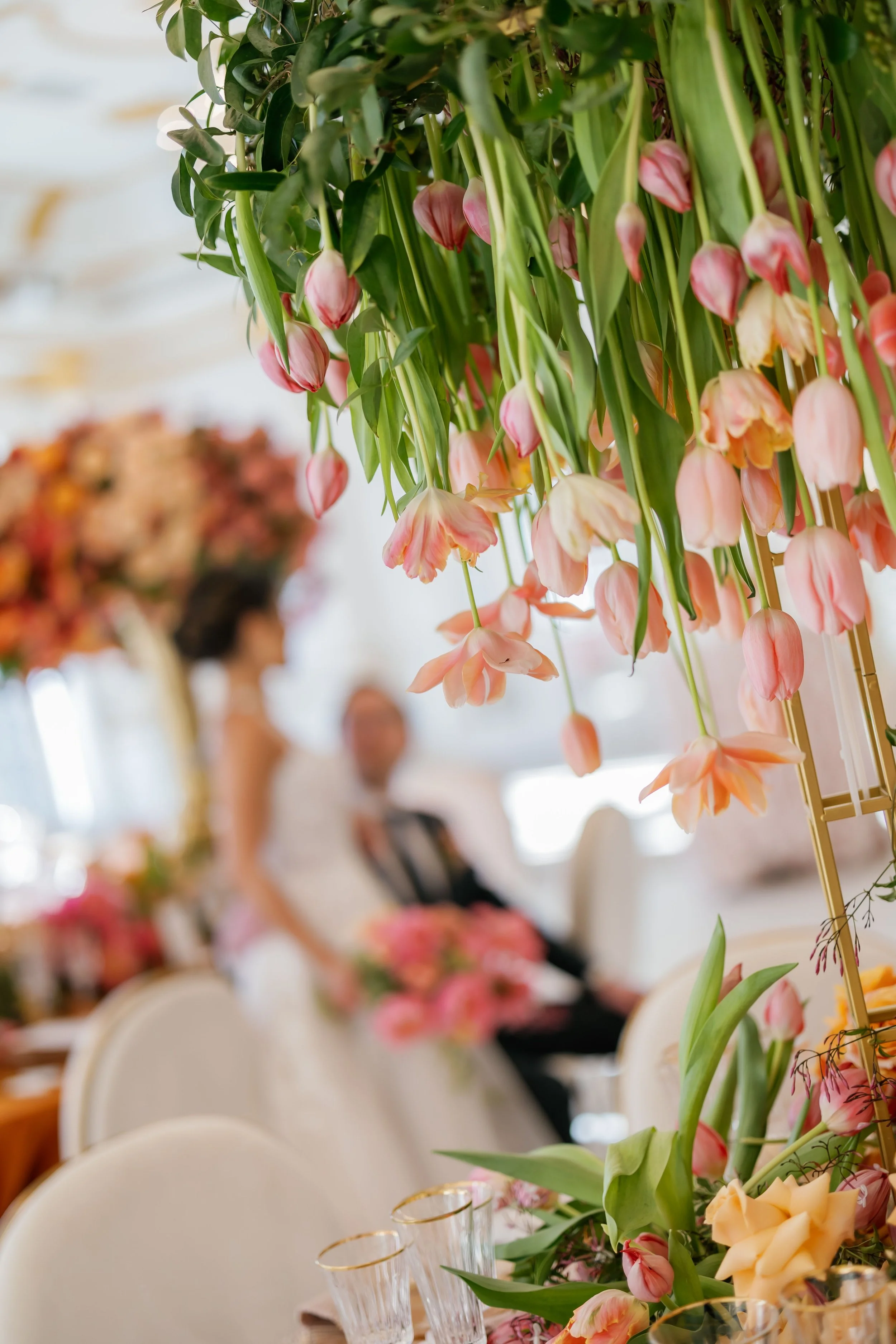 A close-up of hanging pink and cream tulips with green leaves, decorated for a celebration or event, with a blurred background showing people seated at tables.