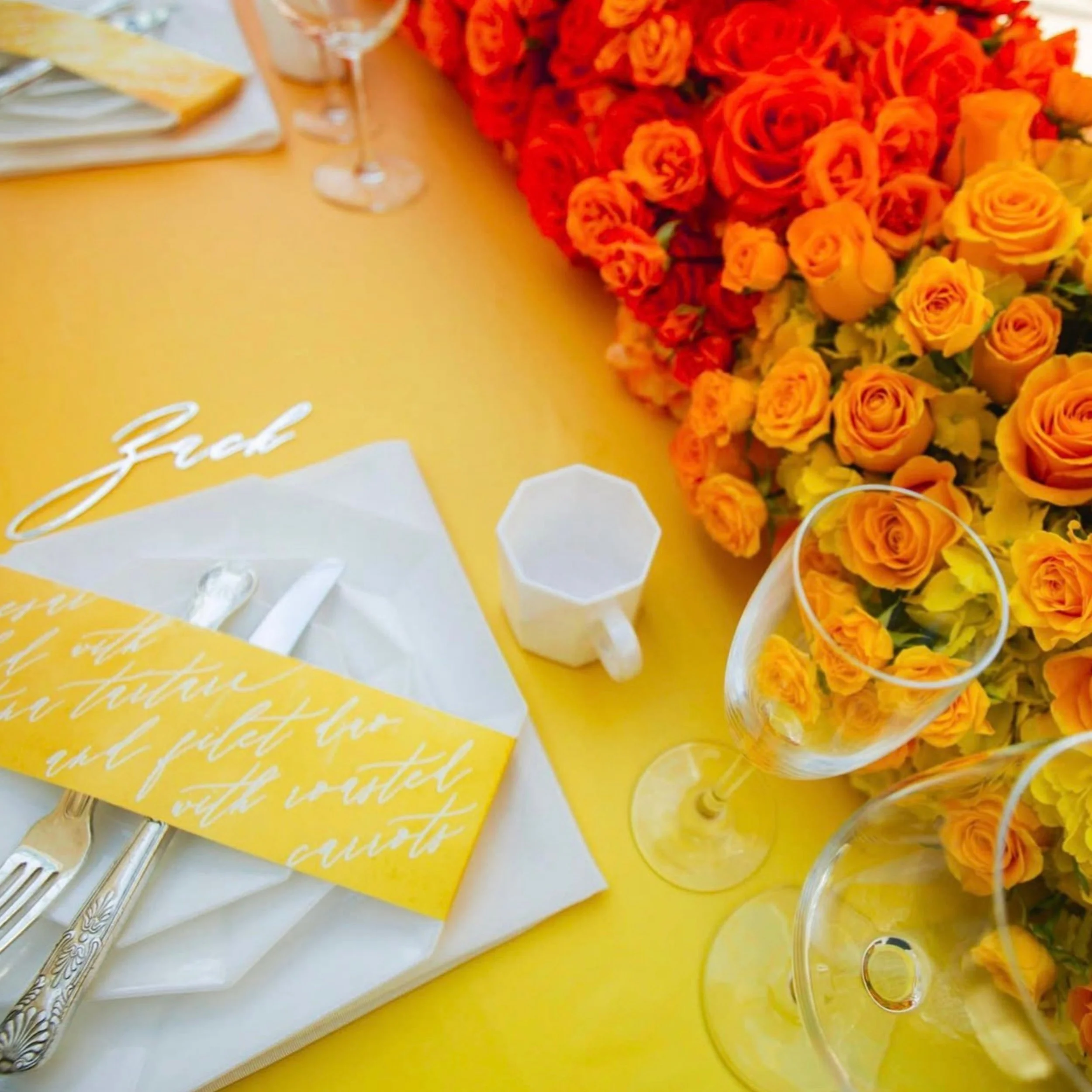 Bright yellow table with a large tablescape of orange and red roses on the right side, a glass of wine, and a stack of white napkins with a yellow cloth underneath.