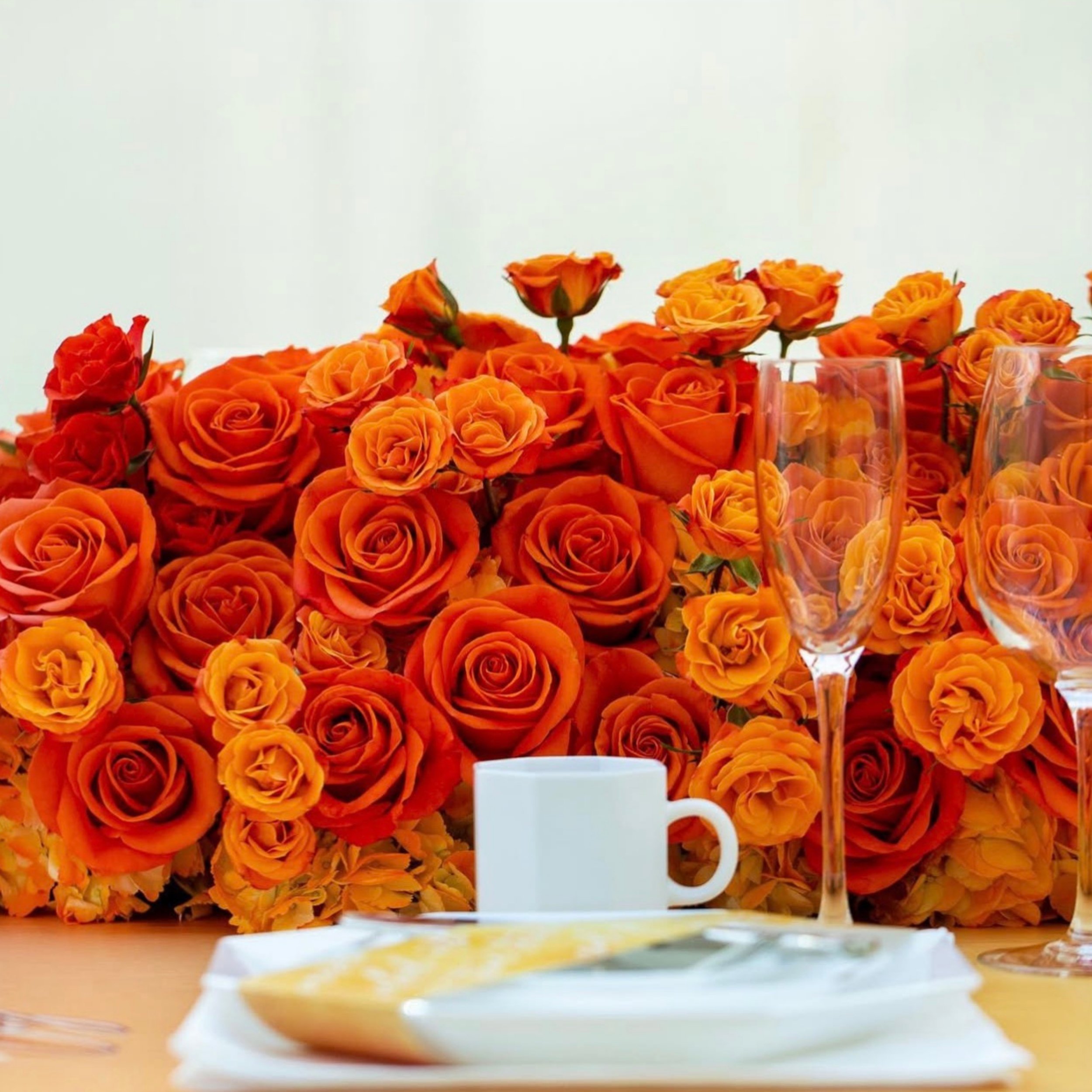 A table decorated with a large arrangement of orange roses, with glasses and a white cup visible in the foreground.
