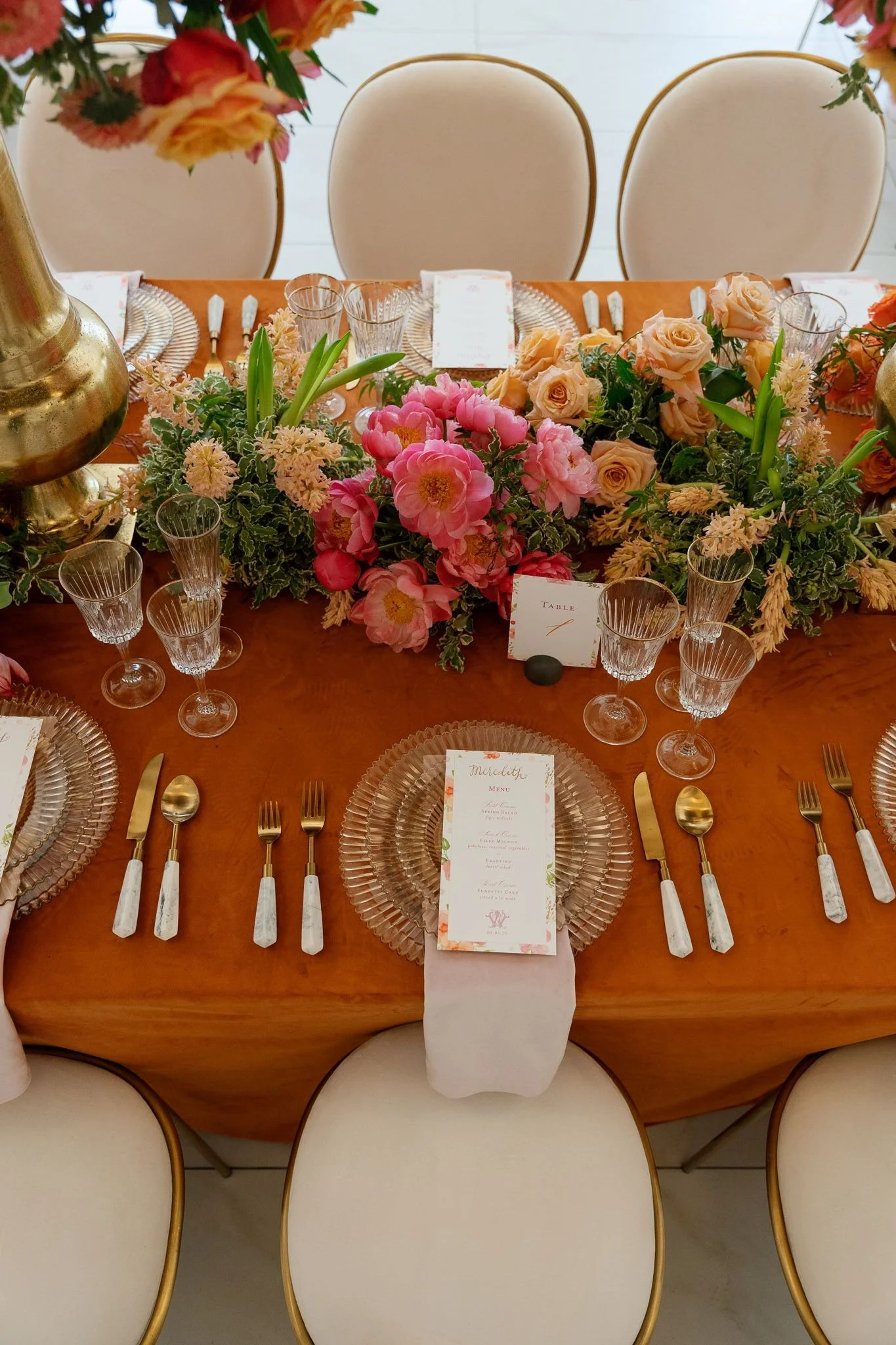 A round wedding or event table set with pink and peach flowers as a centerpiece, gold-rimmed glass chargers, clear glassware, and gold and white flatware. There are five chairs with white cushions and gold trim around the table. A menu card is placed on the table, and a table number card indicates it is table 1.