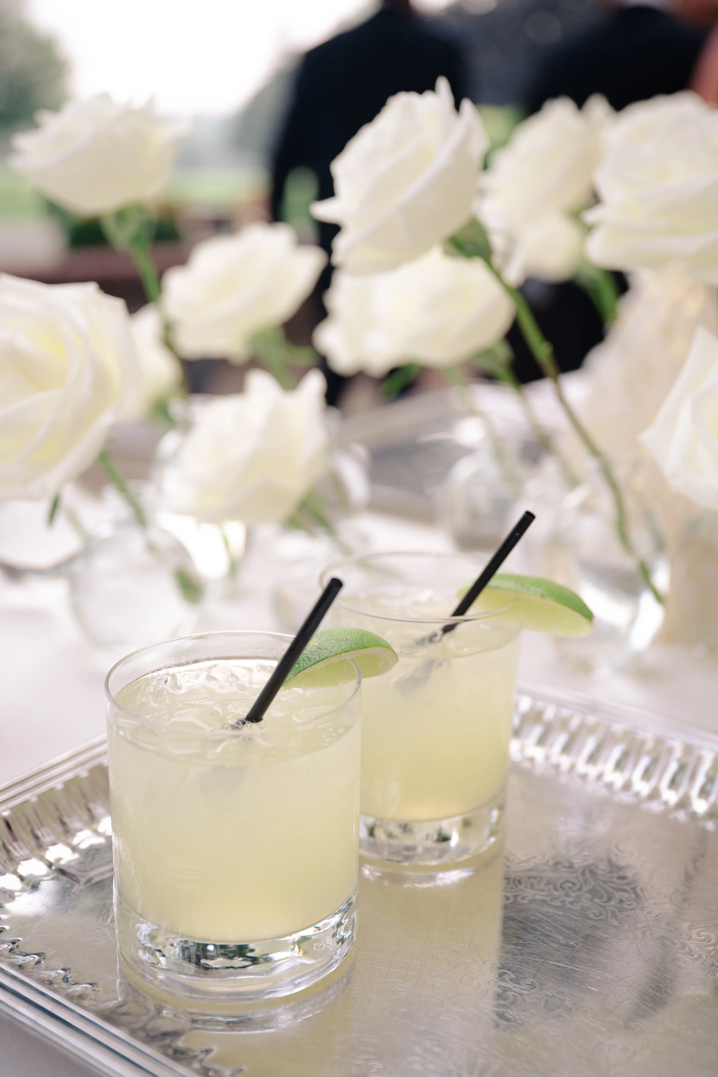 Close-up of three glasses of lemonade garnished with lime wedges and black straws, arranged on a silver tray. In the background, there are white roses, slightly out of focus.