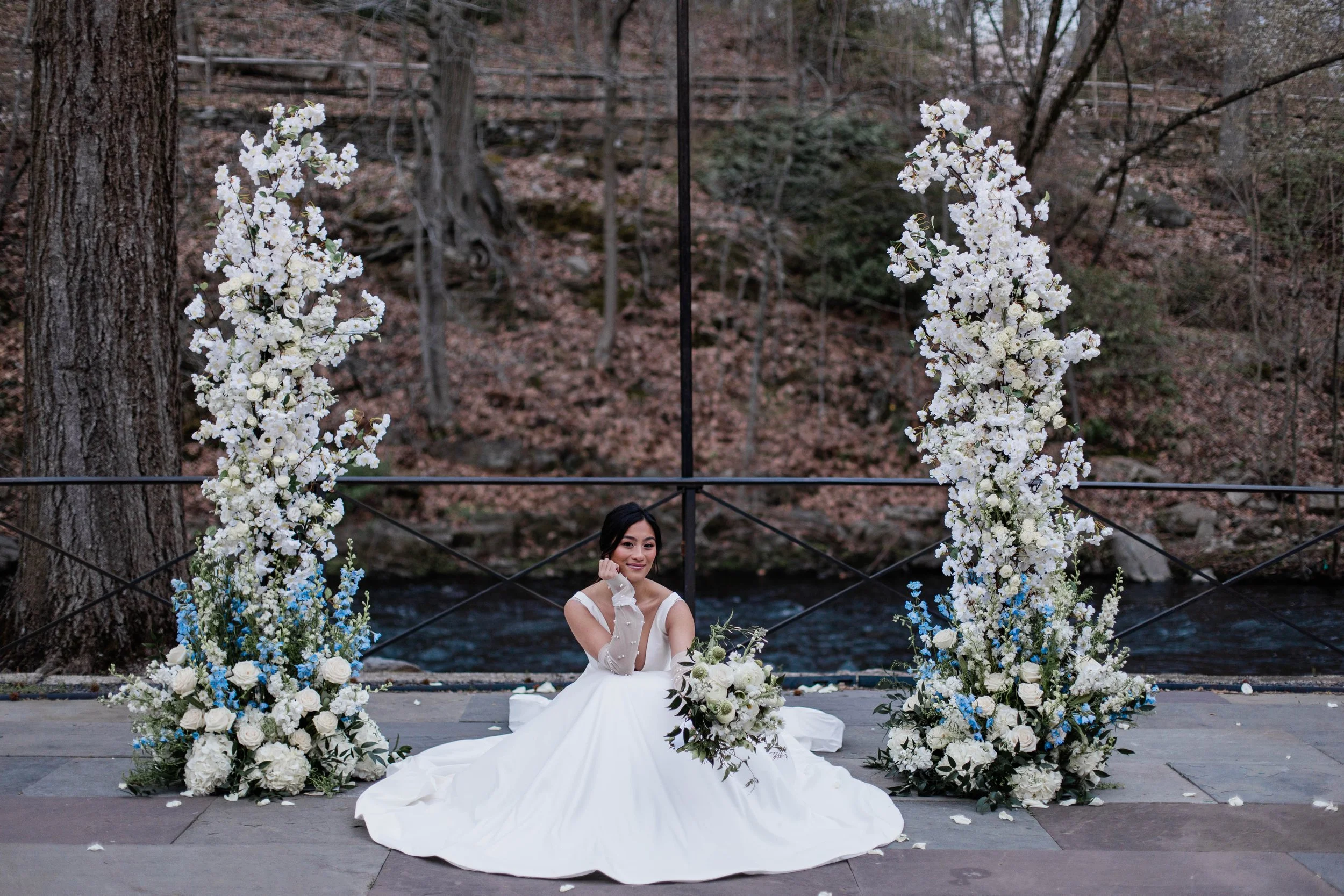 Bride smiling with boquet in her ceremony arch, White florals touched with light blue accents at the base of the broken arch