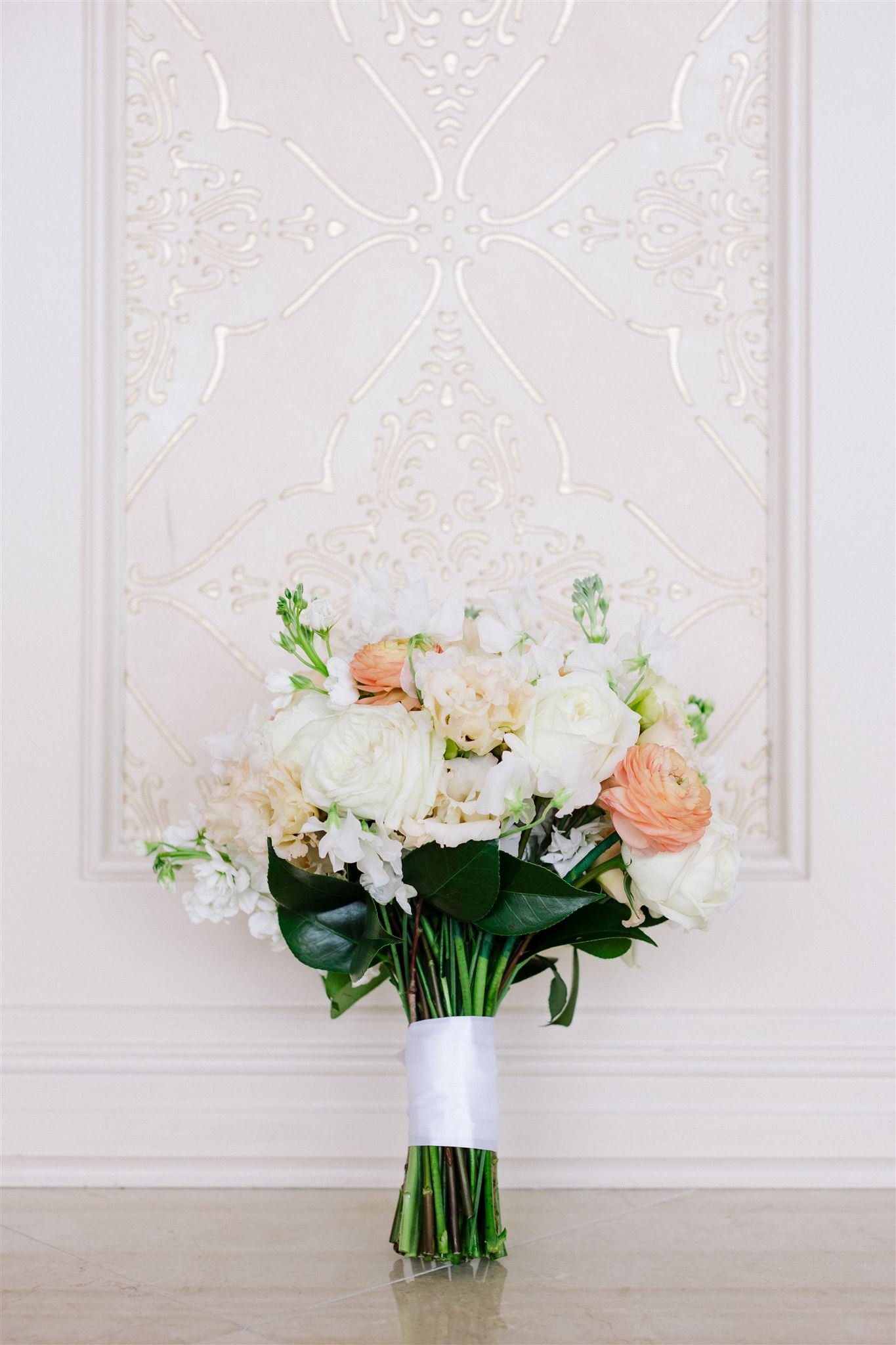 A bouquet of white and peach-colored flowers tied with a white ribbon, standing on a polished floor against a decorative white wall panel.