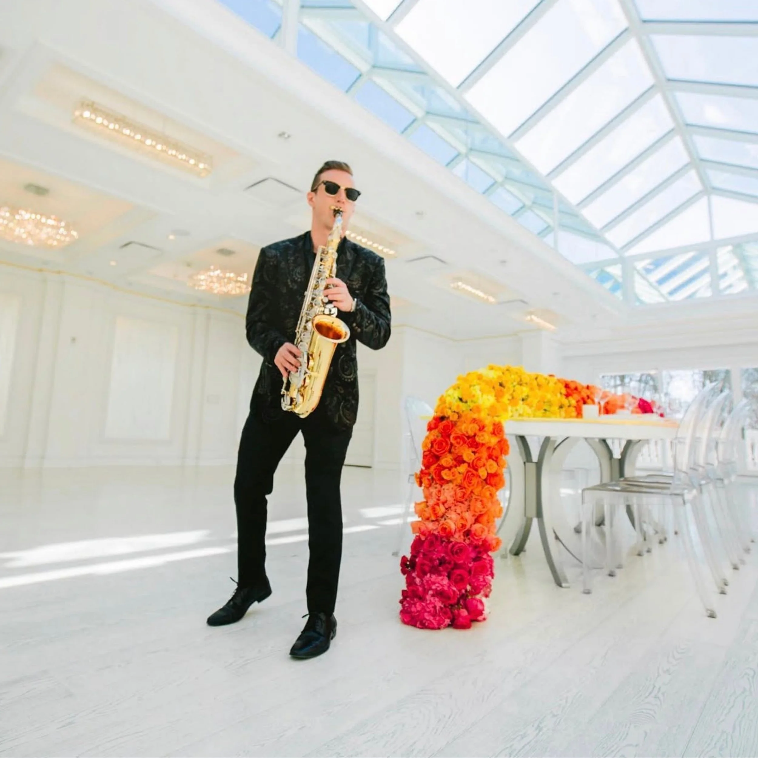 A man in a black suit and sunglasses playing a saxophone indoors near a table with bright orange and pink flower arrangements, with a modern, glass-domed ceiling overhead.
