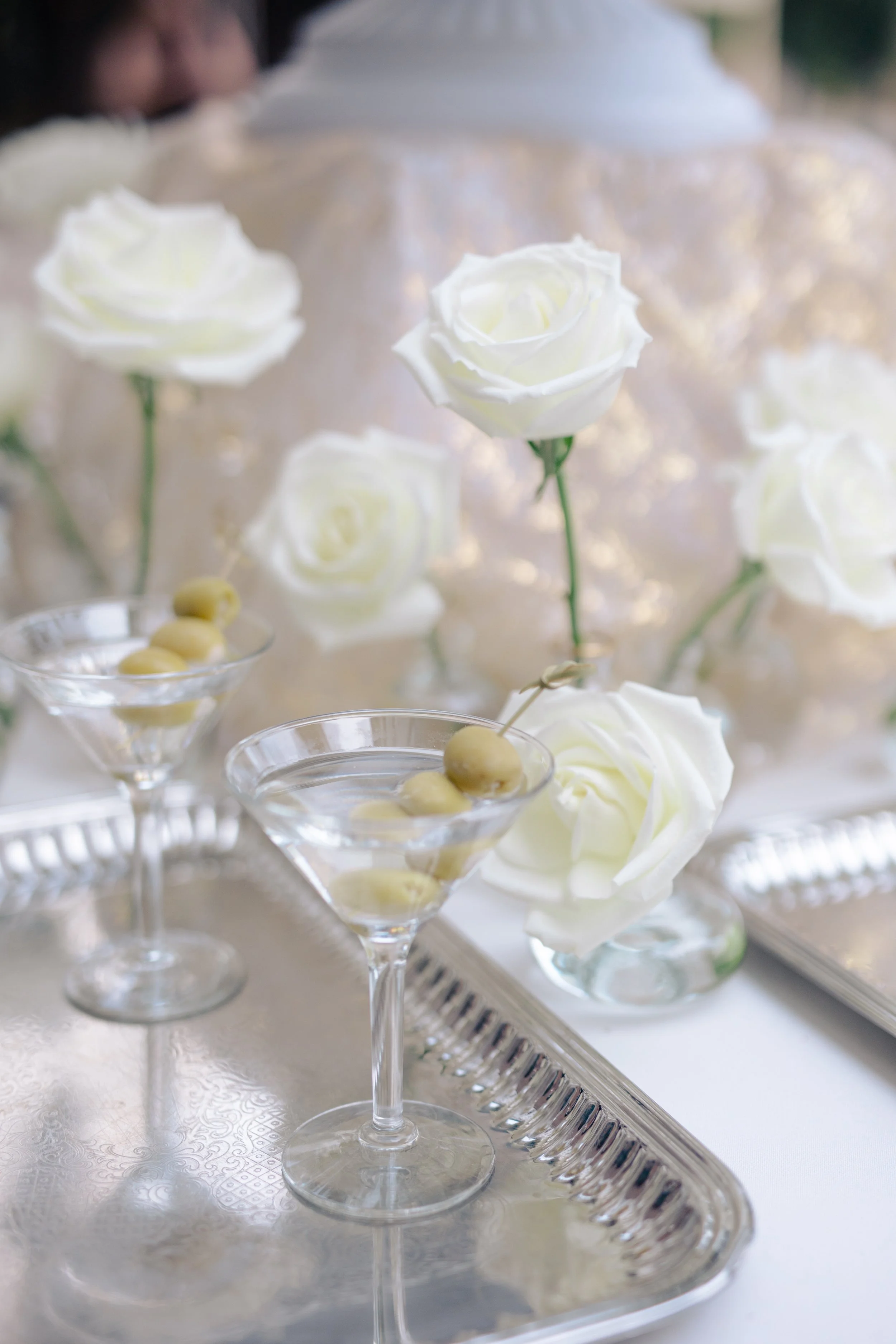 Clear martini glasses filled with white olives, decorated with white roses, arranged on a silver tray and a white tablecloth, in an elegant setting.