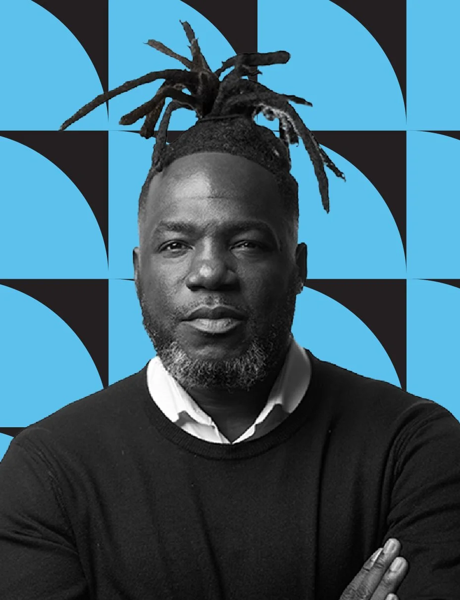 Black man with dreadlocks tied up, wearing a black sweater and white collared shirt, standing in front of a geometric blue and black patterned background.