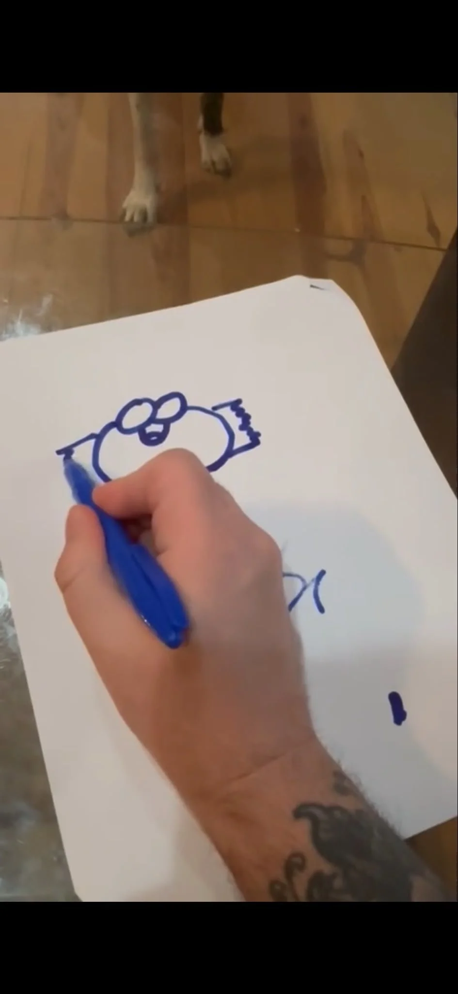 A hand drawing a cartoon character with a blue marker on a sheet of white paper, with a dog standing on a wooden floor in the background.