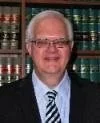 Portrait of a man with glasses and white hair wearing a suit and tie, standing in front of bookshelves.