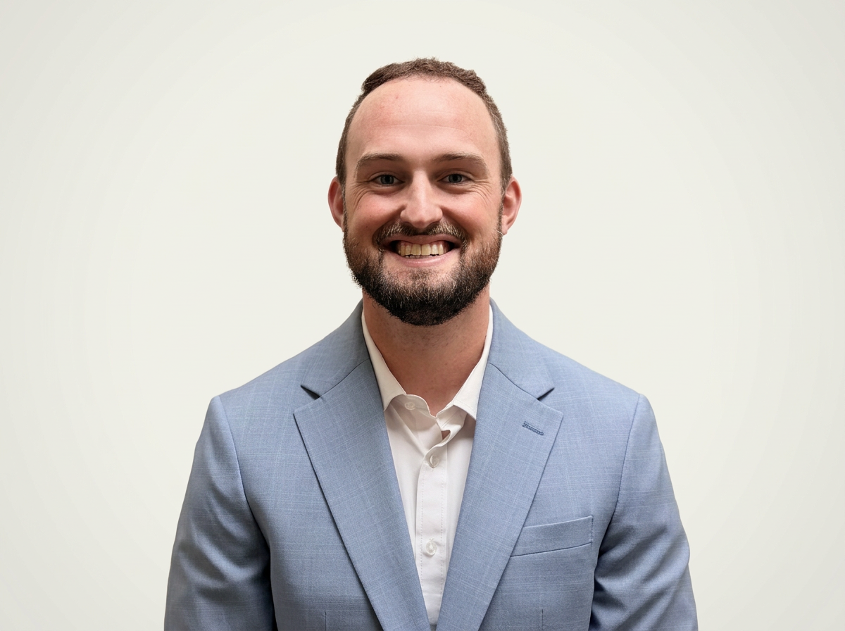 Portrait of a smiling man with a beard wearing a light blue suit jacket and white shirt against a plain light background.