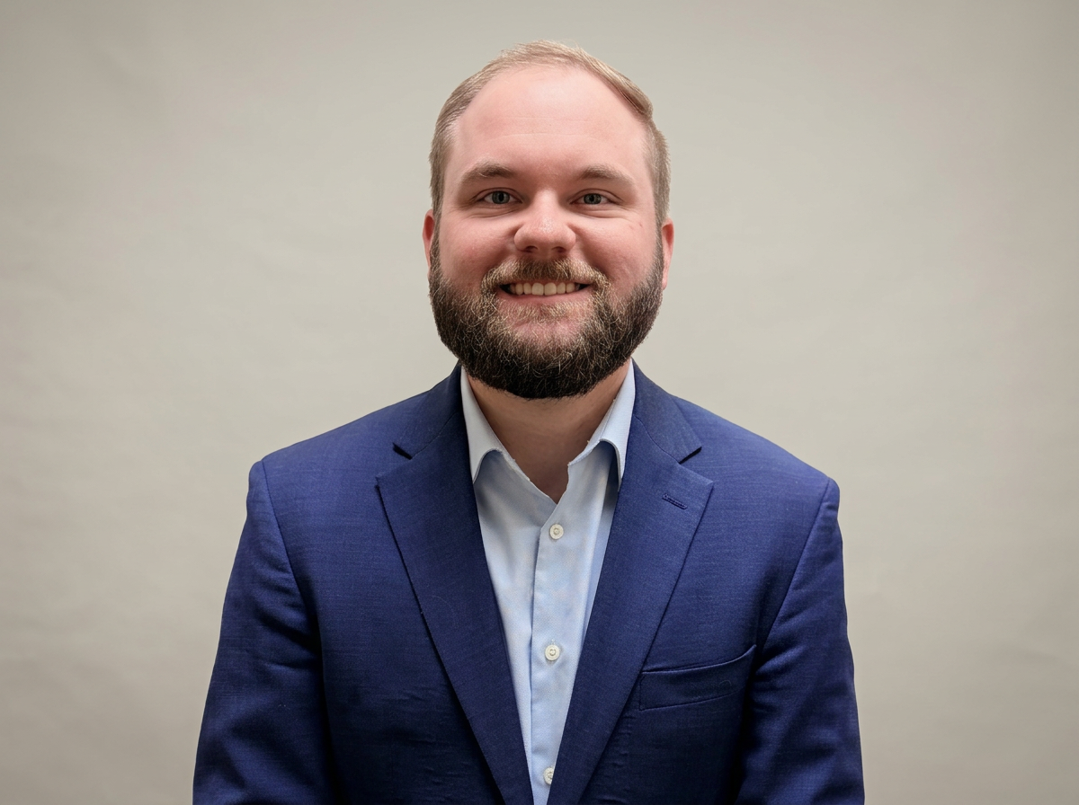 A smiling man with a beard in a blue suit and light blue shirt, standing against a plain beige background.