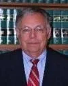 A man with glasses and gray hair wearing a suit and red tie, standing in front of a bookshelf filled with books.