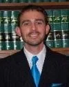 Portrait of a man in front of a bookshelf filled with legal or reference books, wearing a dark suit, white shirt, and blue tie.