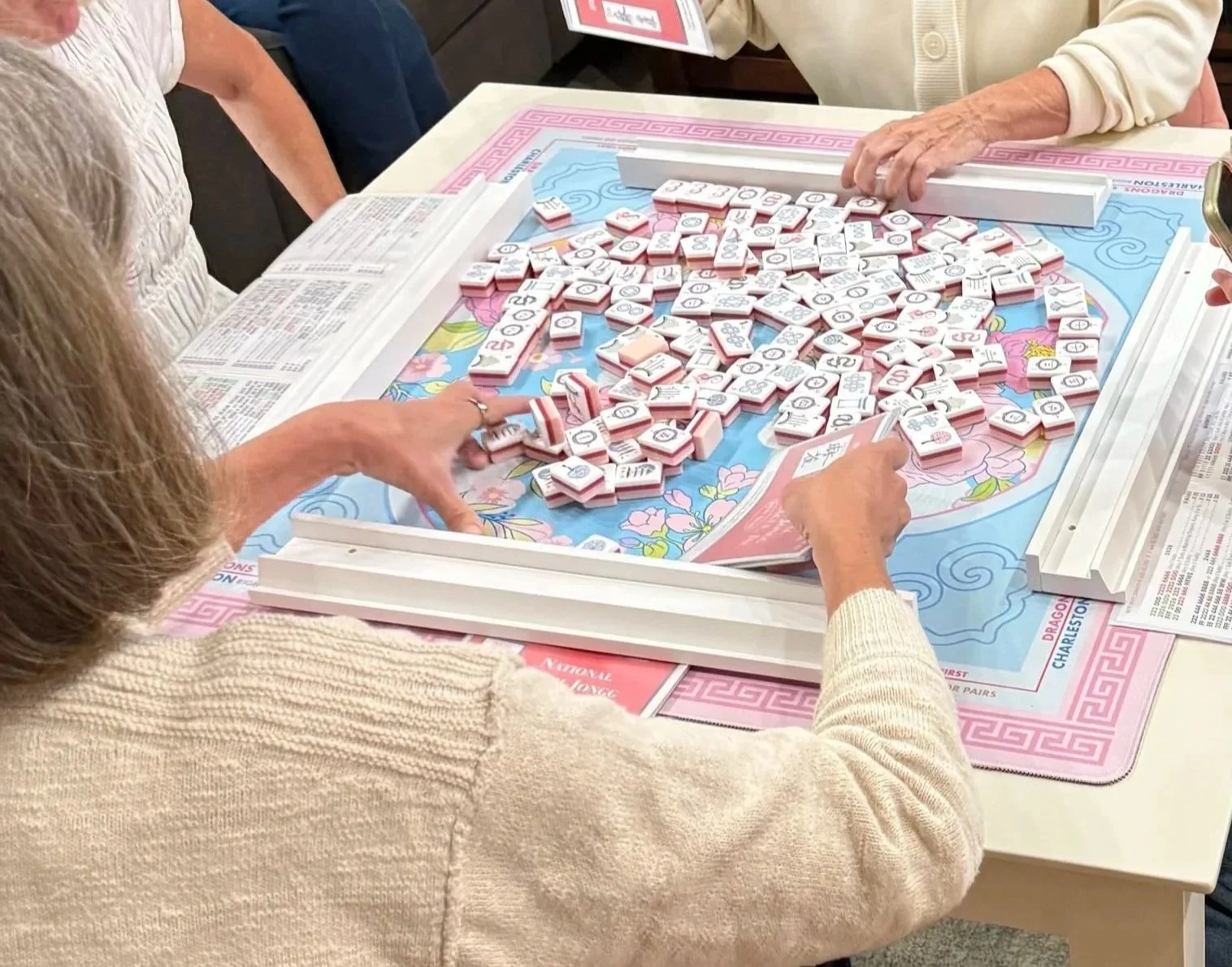 Several women playing a game of Mahjong around a table, with Mahjong tiles scattered across the game board.