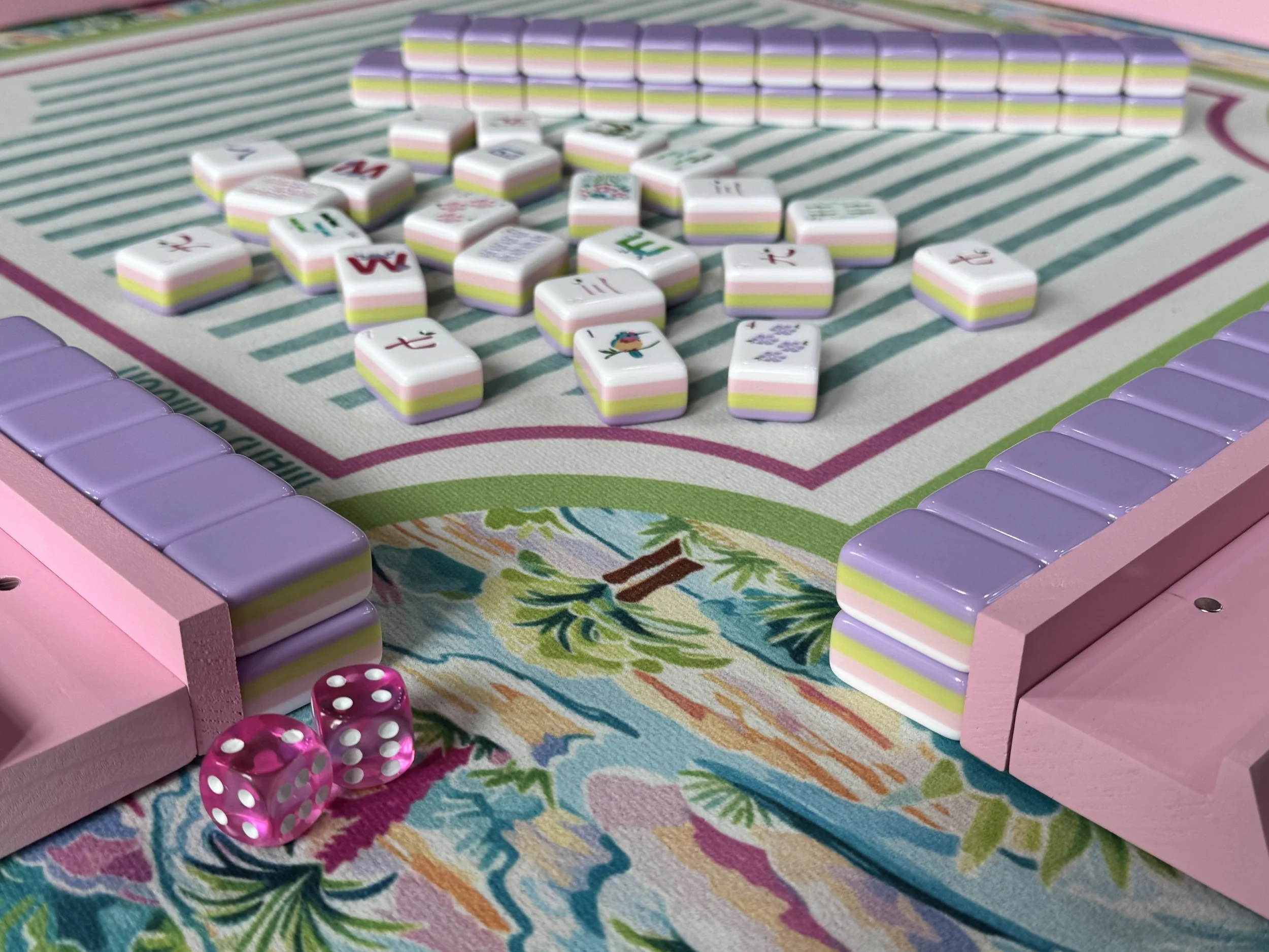 Colorful board game with pink, purple, green, white, and yellow pieces, along with pink dice, on a decorative fabric surface.