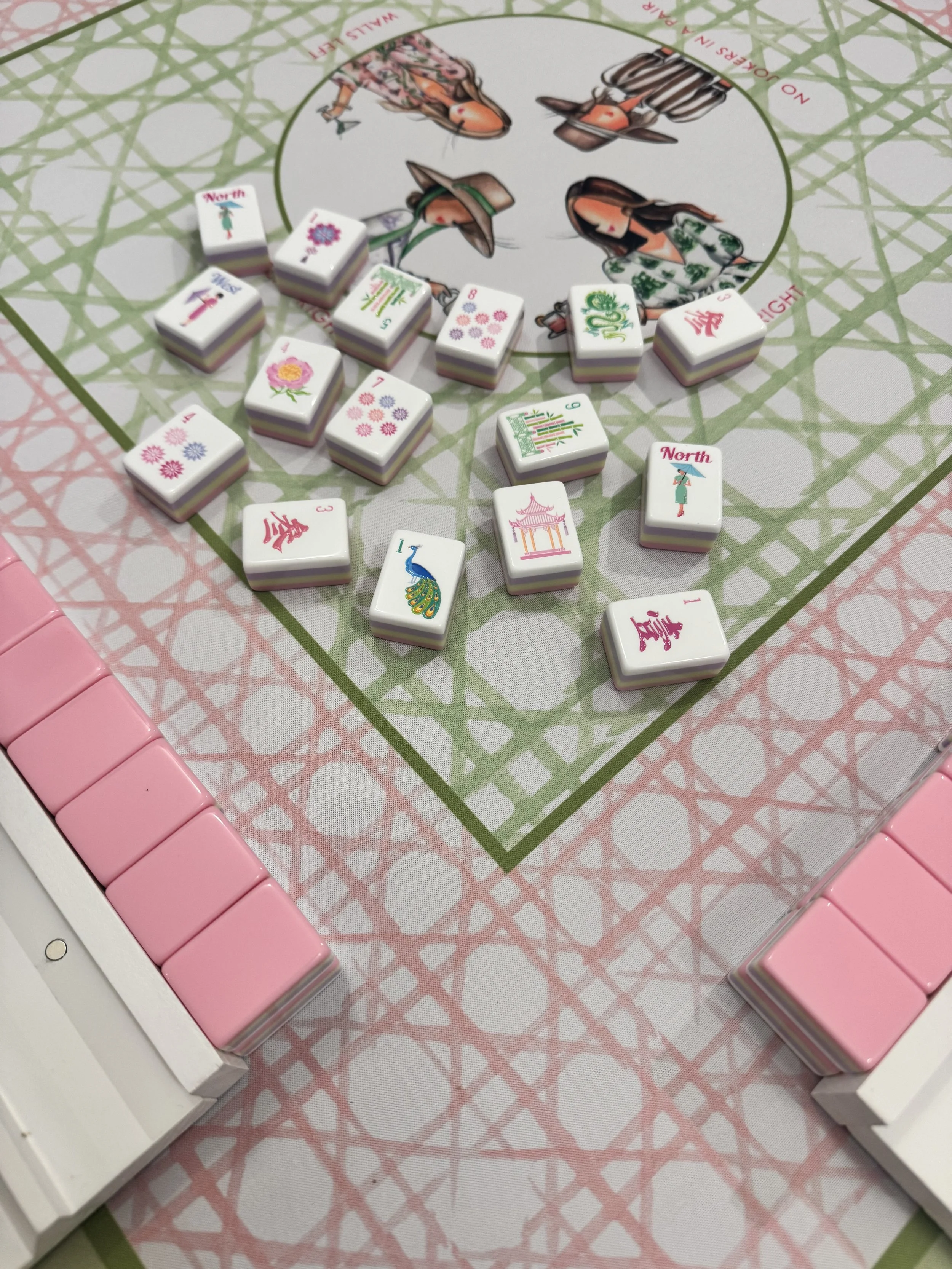 A close-up view of a Mahjong or domino game setup featuring multiple white tiles with colorful illustrations, set on a decorative game mat with illustrations of women in traditional Asian attire, surrounded by pink and green borders.