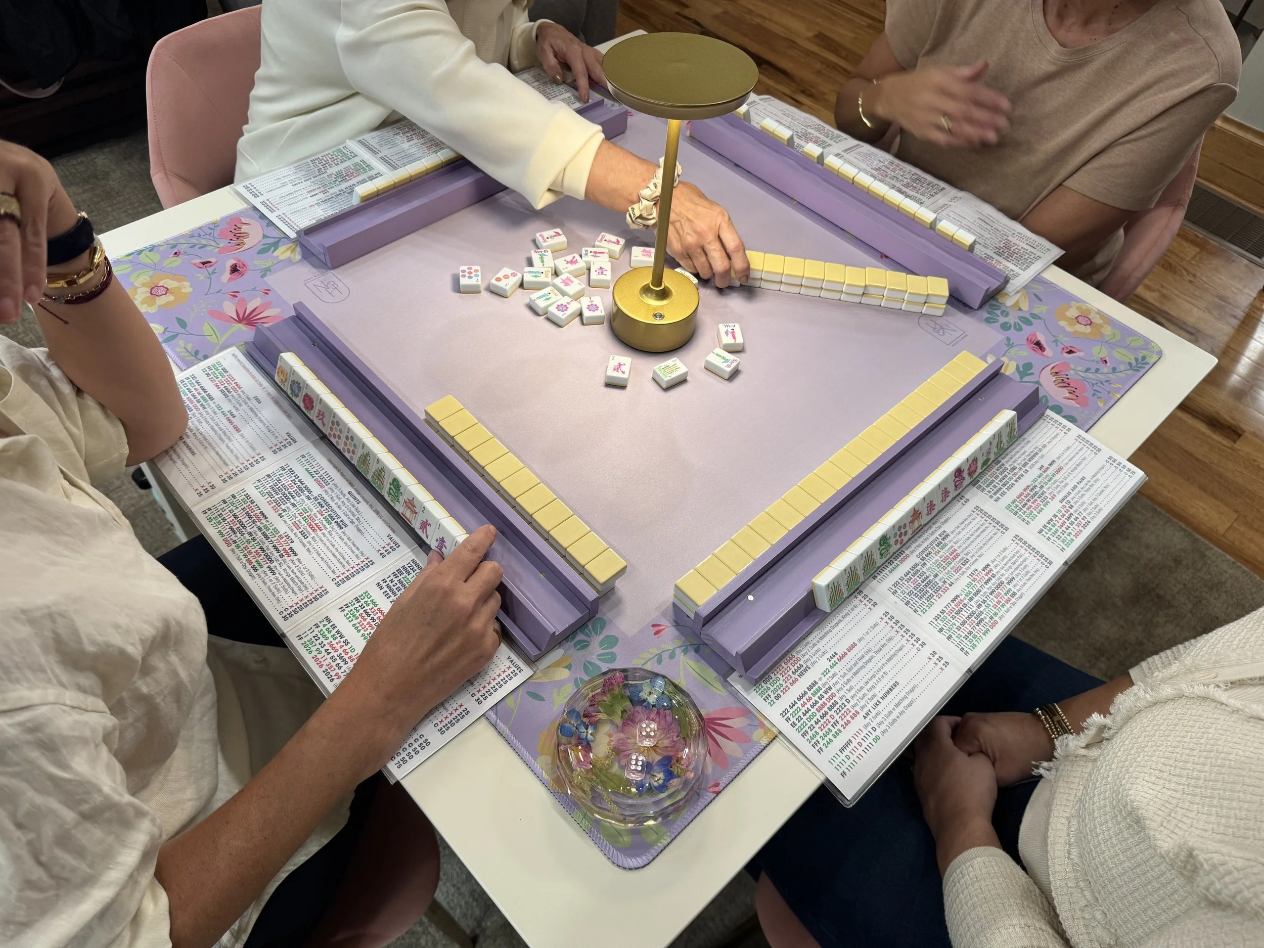 People playing mahjong around a table with a purple tablecloth, mahjong tiles, and a bowl of decorative flowers.