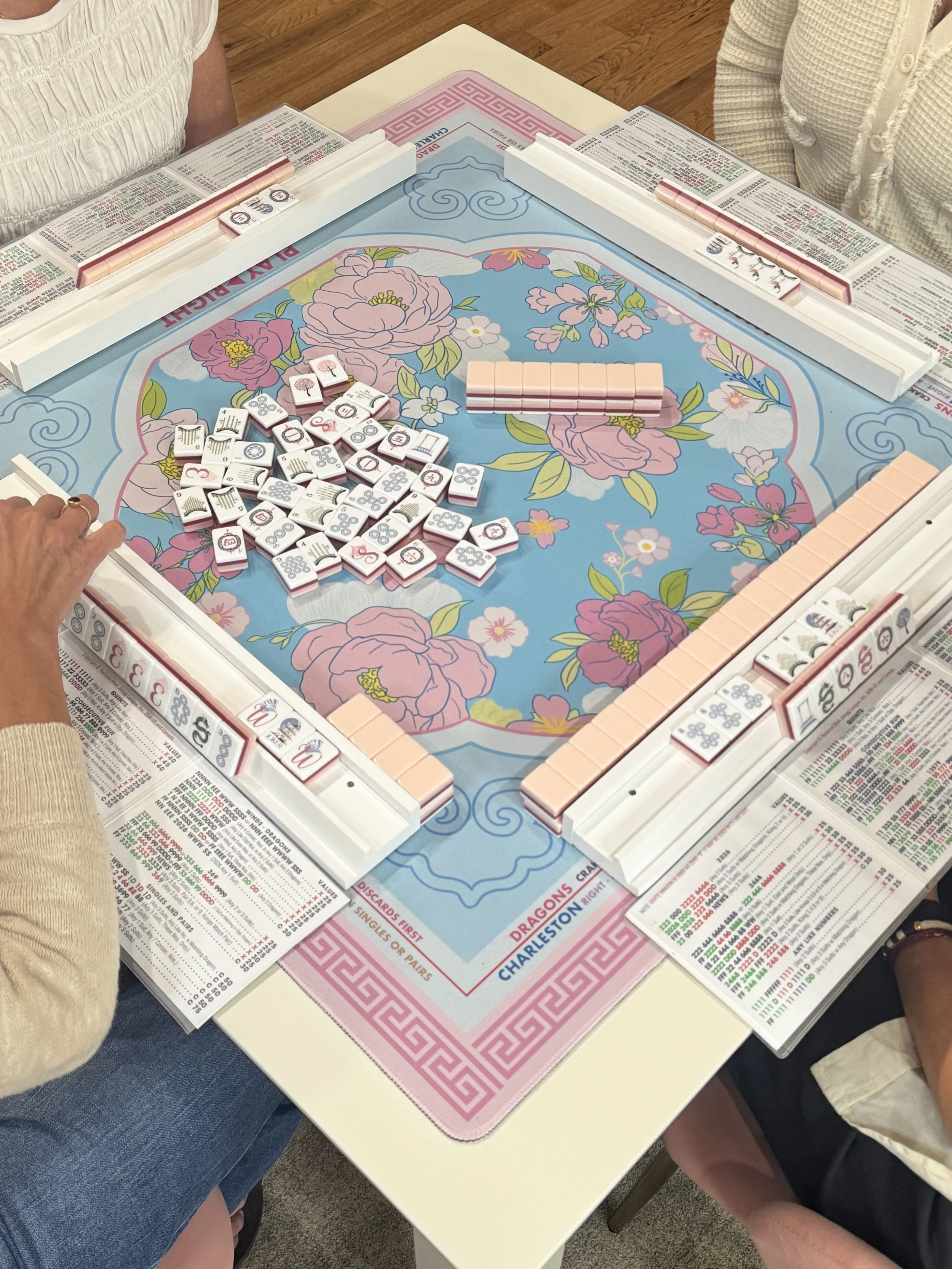 People playing Mahjong at a table with colorful floral patterns on the playing surface. Mahjong tiles are arranged on the table, and players have reference sheets with game rules and scoring.