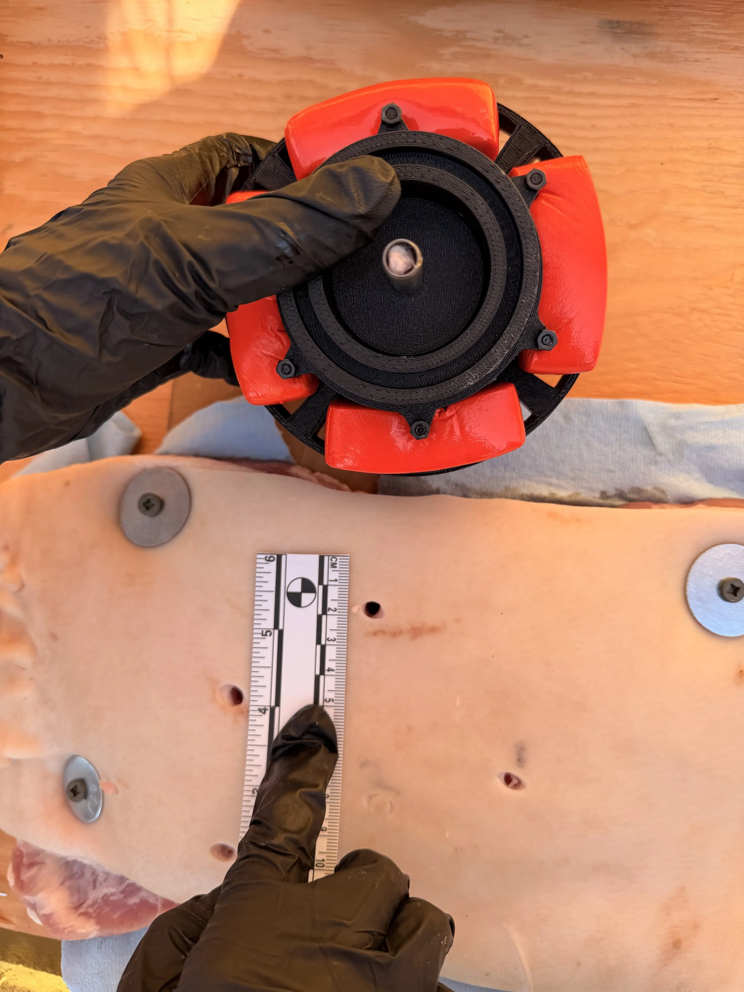 Top-down view of a person wearing black gloves and black shoes, holding a black device with a red portion, over a wooden surface with metal screws and a measurement ruler.