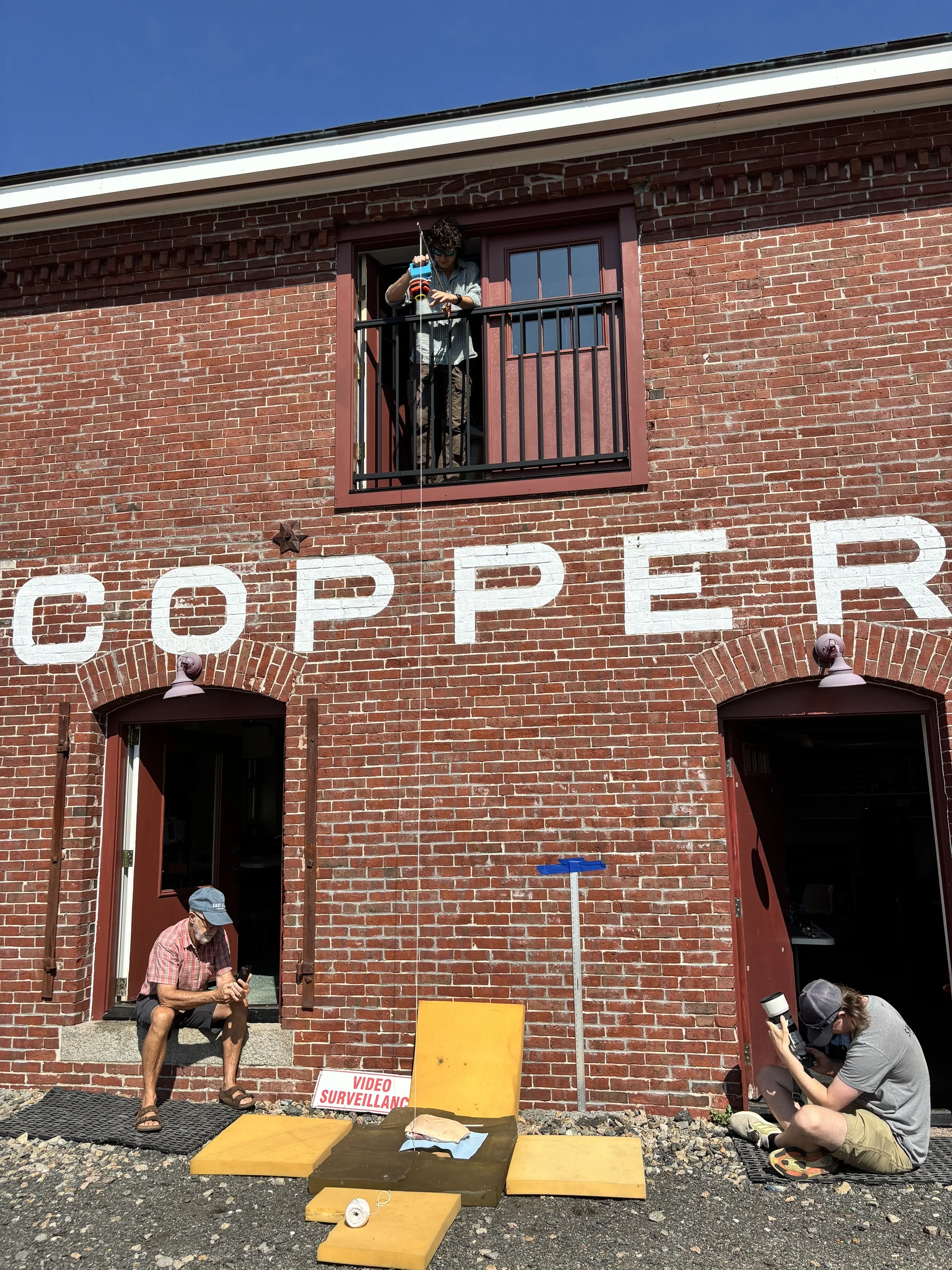 People sitting outside a brick building with the word 'CORNER' partially visible, engaged in photography and videography, with a sign reading 'VIDEO SURVEILLANCE' on the ground.