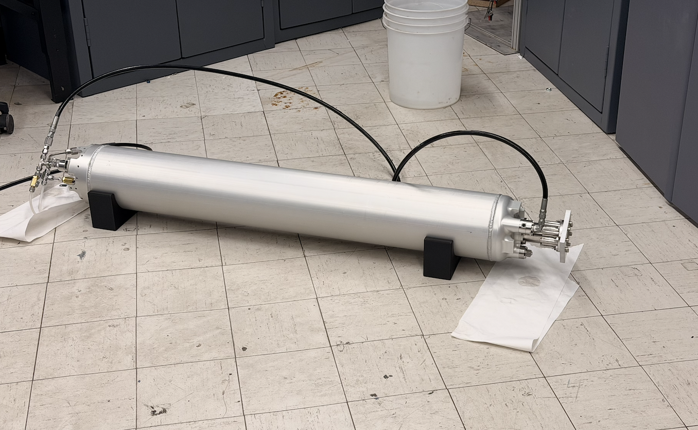 A large metallic cylindrical vacuum tube with multiple fittings and cables, resting on the tiled floor of a laboratory or workshop, supported by black foam blocks.