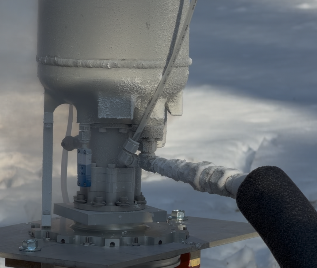 Close-up of a weather station with frost, showing a pole and instrument with snow-covered ground and cloudy sky in the background.