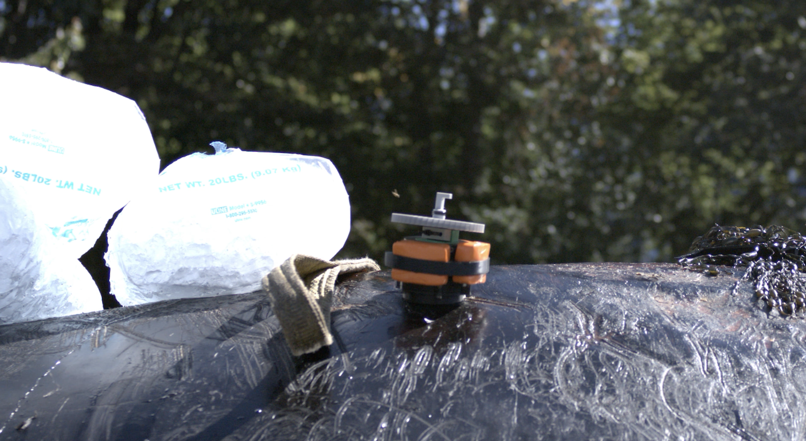 Close-up of a tree trunk with a small, orange and black drone resting on it, two white bags labeled 'Net Wt. 20 lbs,' and a chain on the right. Blurred green trees in the background.