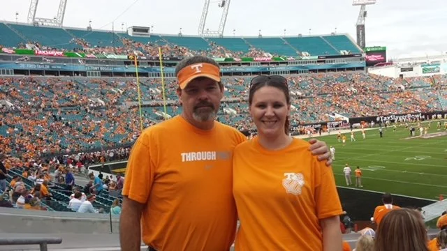 Two people wearing orange shirts at a football stadium, with fans and the field in the background.