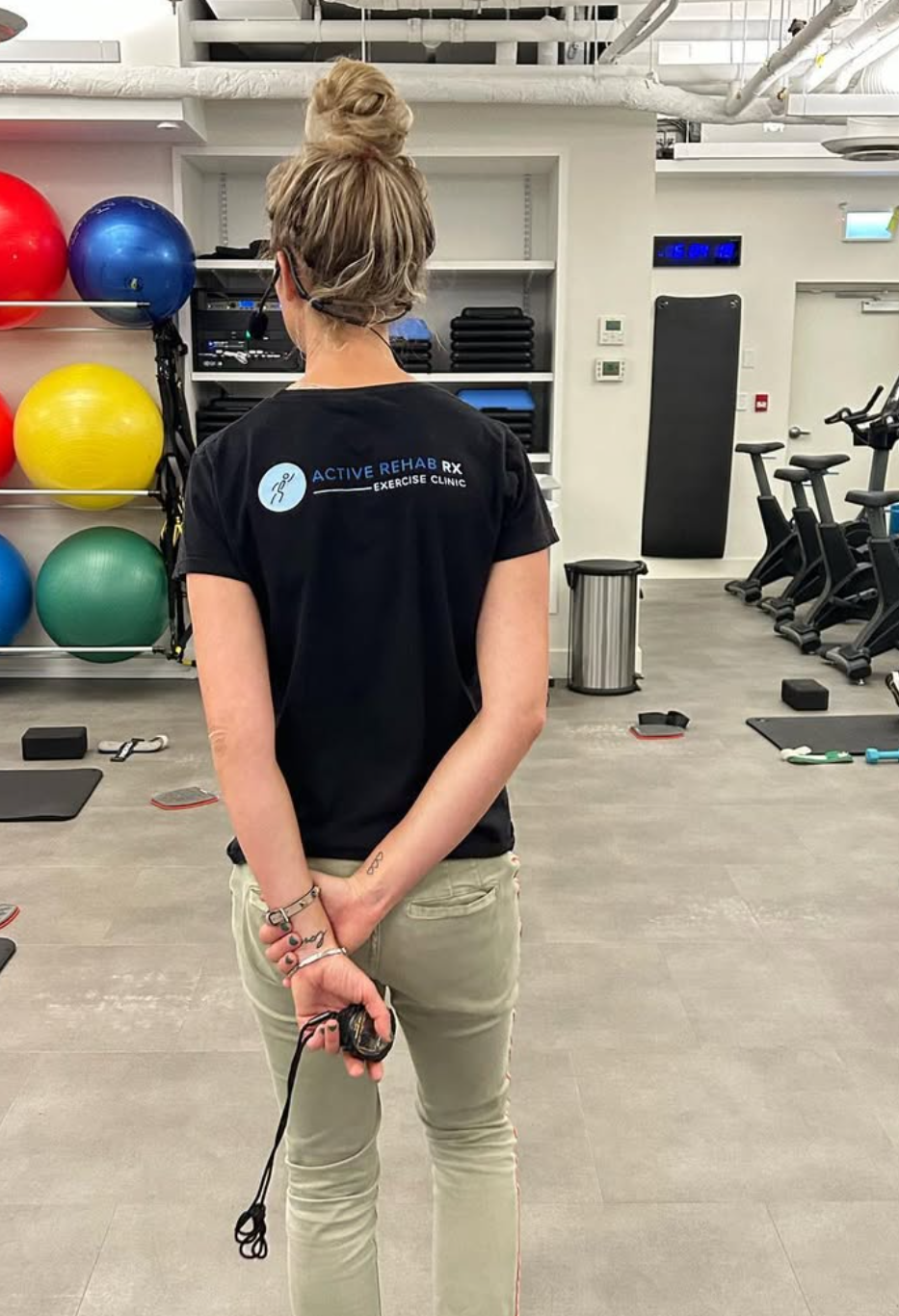 A person with a bun hairstyle holding a jump rope behind their back, standing in a gym with exercise equipment and colorful stability balls.