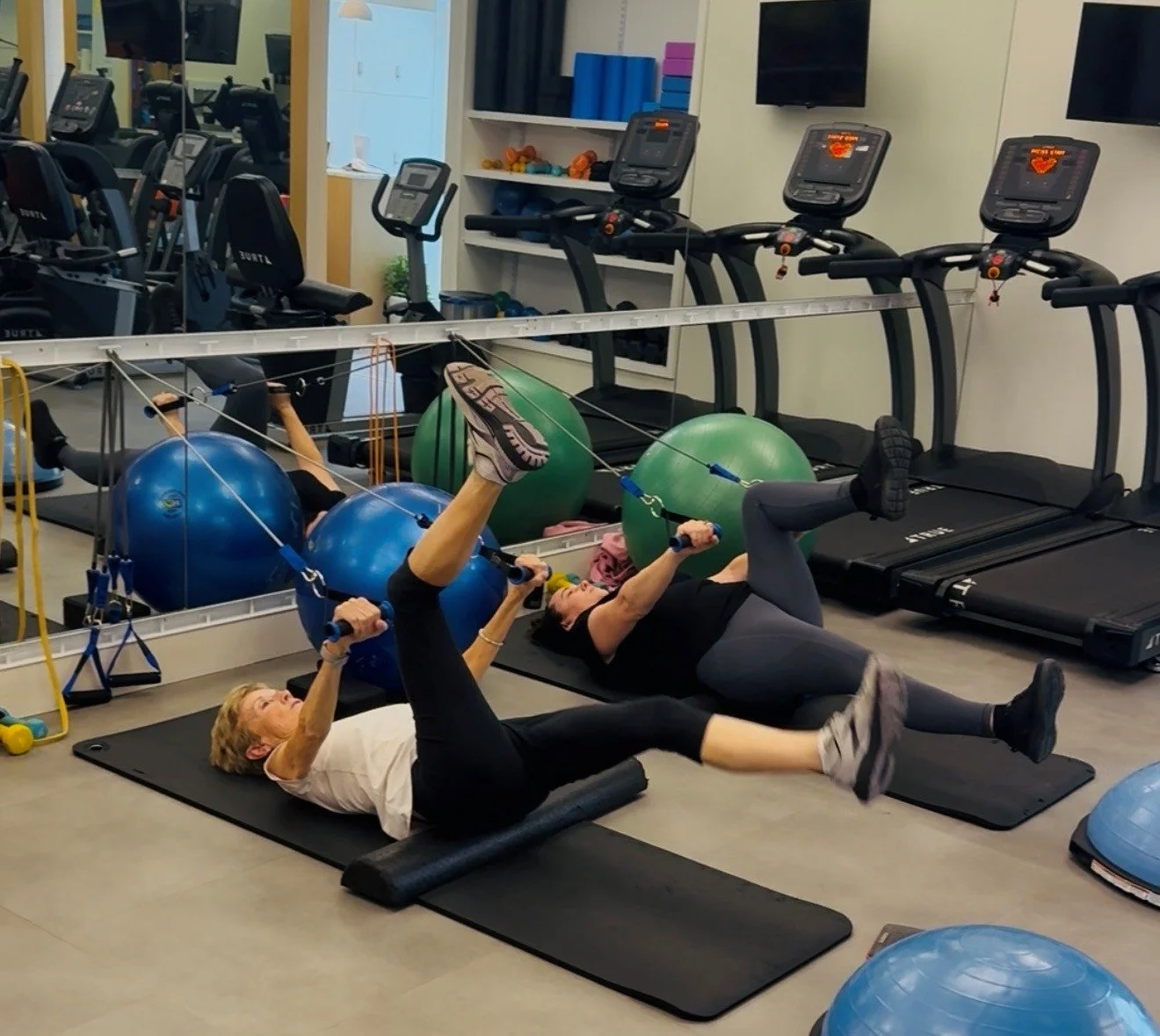 Two women exercising on mats in a gym, using resistance bands attached to exercise balls as part of their workout.