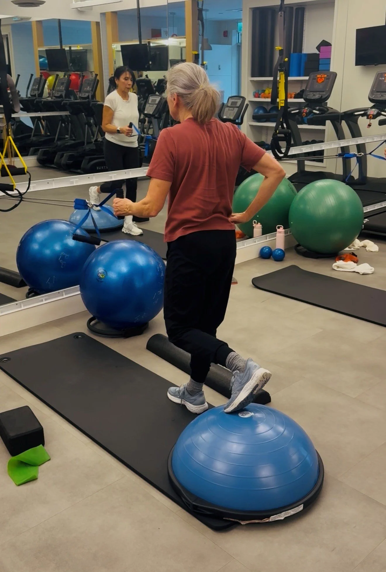 A woman doing step exercises on aerobic step platforms in a gym, with large exercise balls and mirrors. Another woman is in the background, exercising with resistance bands.