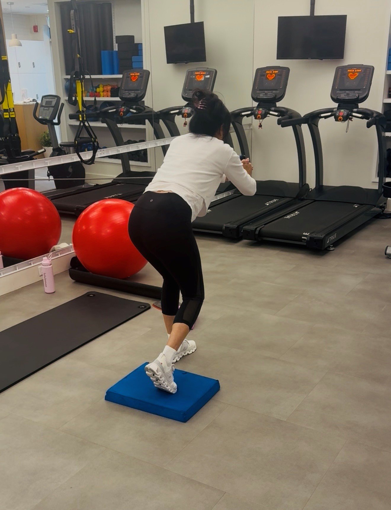 A woman exercising on a step platform in a gym with treadmills, red exercise balls, and workout equipment.