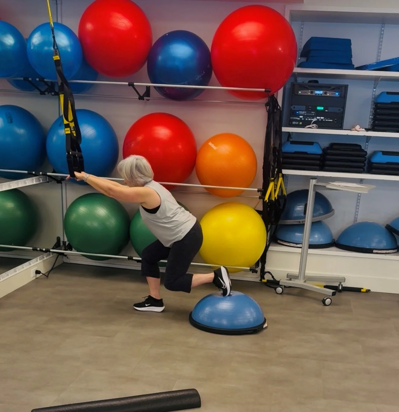 Older woman exercising with resistance bands attached to a wall in a fitness studio with colorful stability balls and exercise equipment.