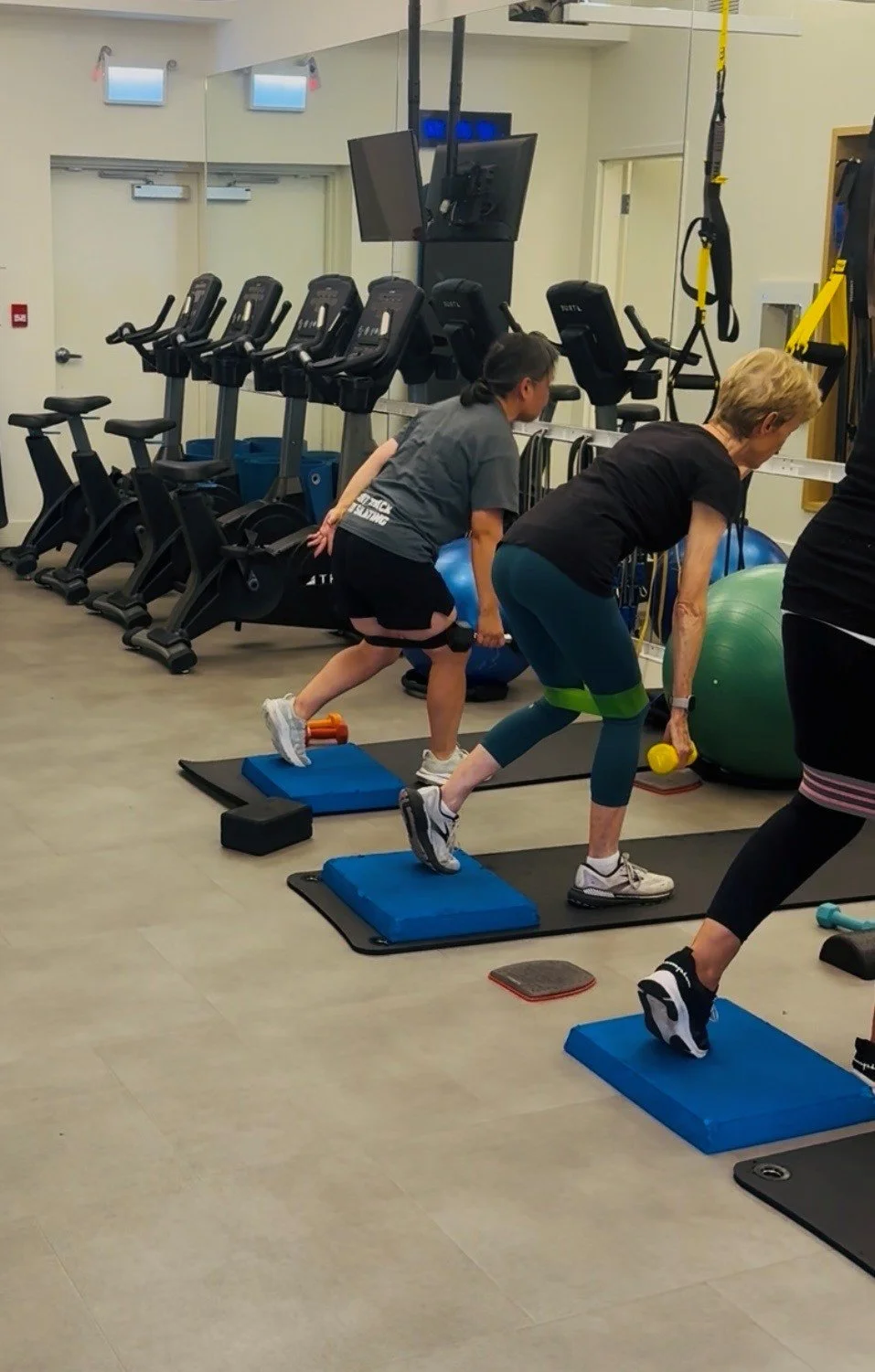 Two women in fitness classes using hand weights while performing leg exercises on blue padded mats in a gym with cardio equipment and exercise balls.