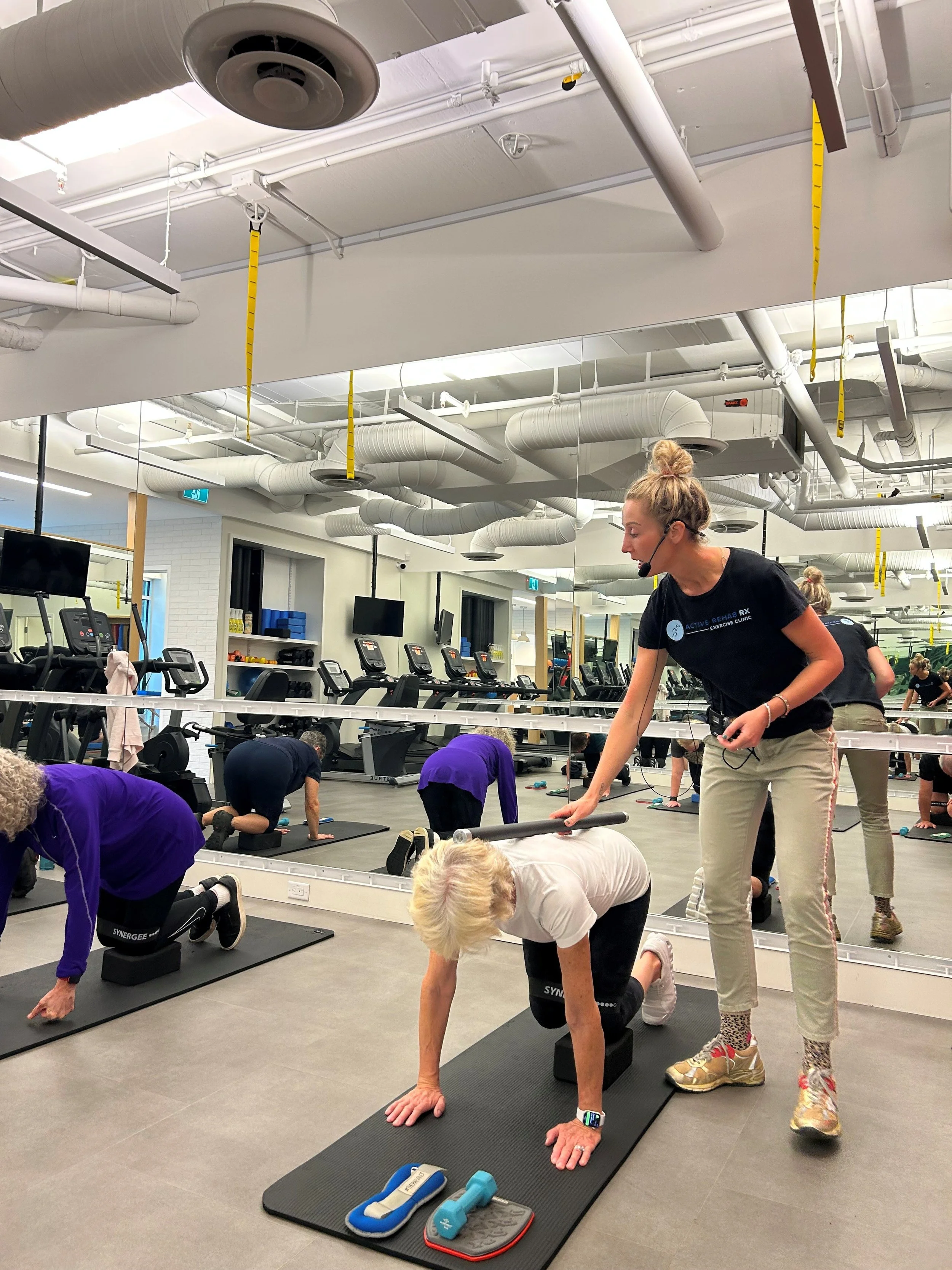 A group exercise class inside a gym with a mirrored wall, treadmill machines, and exposed ductwork on the ceiling. People are performing yoga poses on mats, with a woman in a black shirt assisting an older woman in a white shirt, both in a downward dog position. Yoga equipment such as blocks and water bottles are visible on the mats.