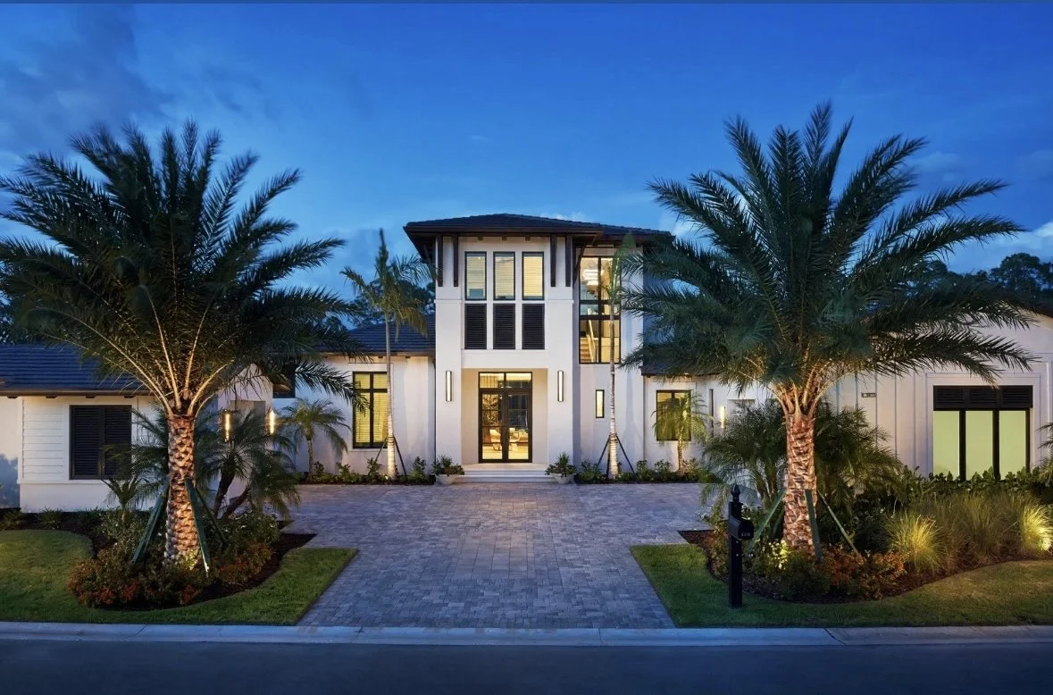 Exterior view of a modern house at dusk with palm trees, well-lit entryway, and a paved driveway.