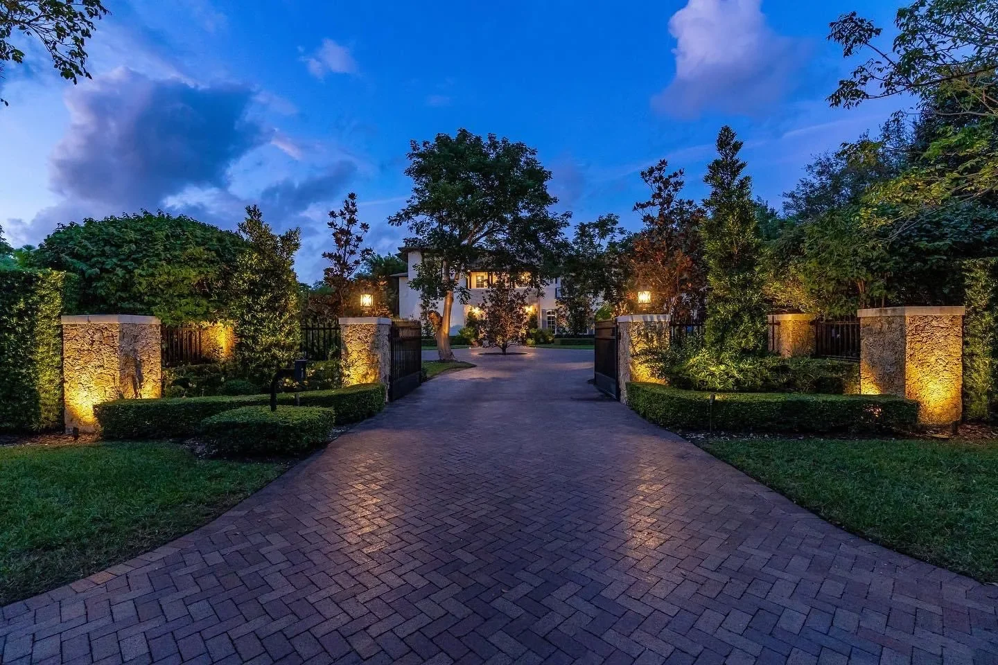A driveway lined with lit landscape lighting leading up to a house at dusk, with trees and bushes on either side and a cloudy sky overhead.