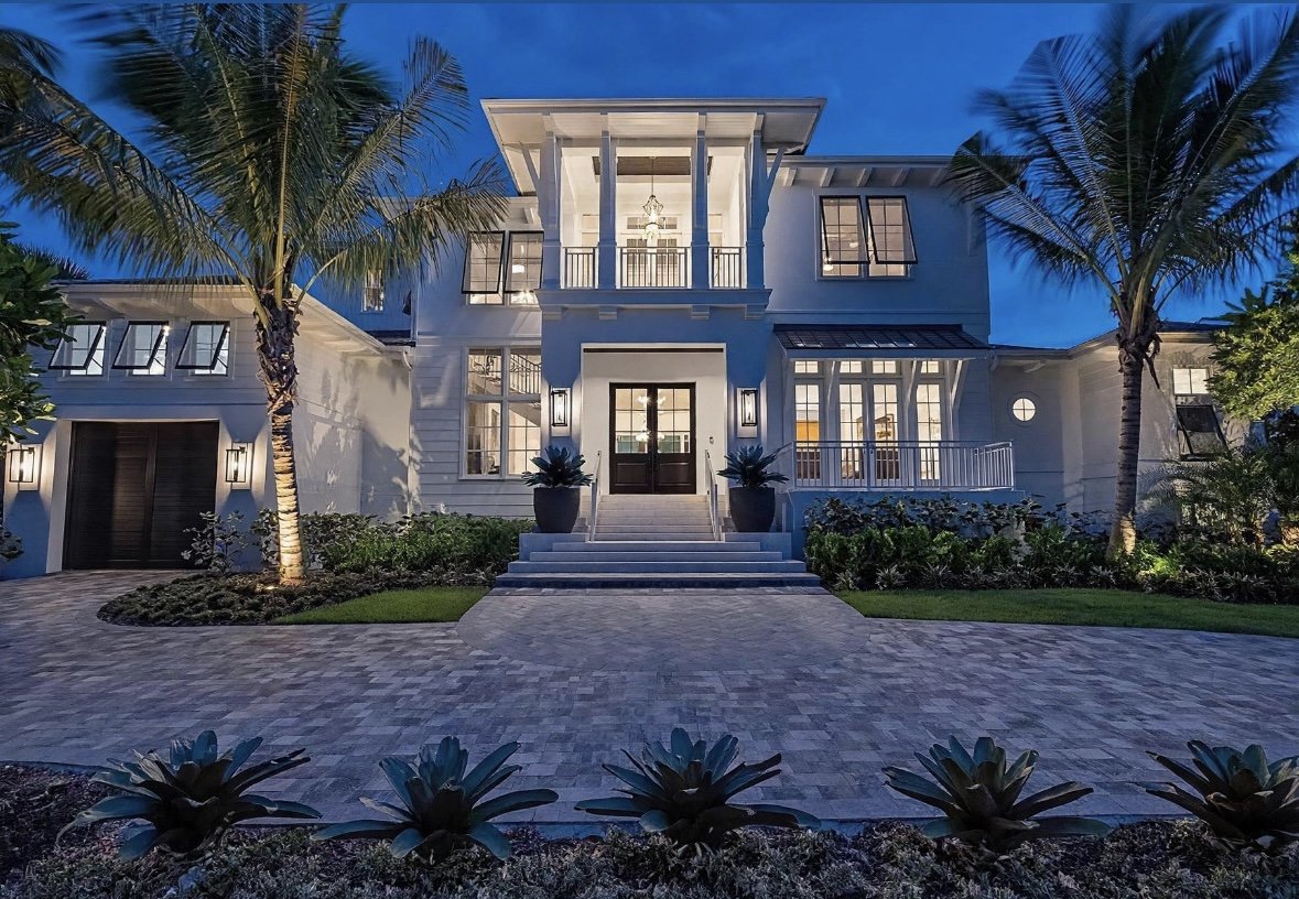 A large, modern white house with a bright exterior, front steps, and large glass windows, illuminated at dusk with palm trees and landscaping in the yard.