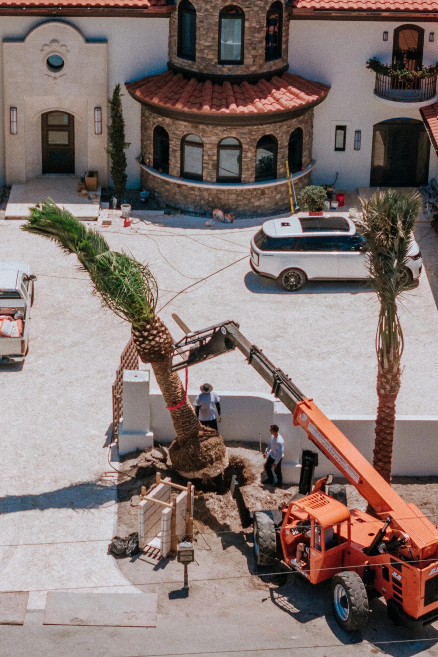 Construction site with workers, palm trees, and a house with a rounded tower and red tile roof.