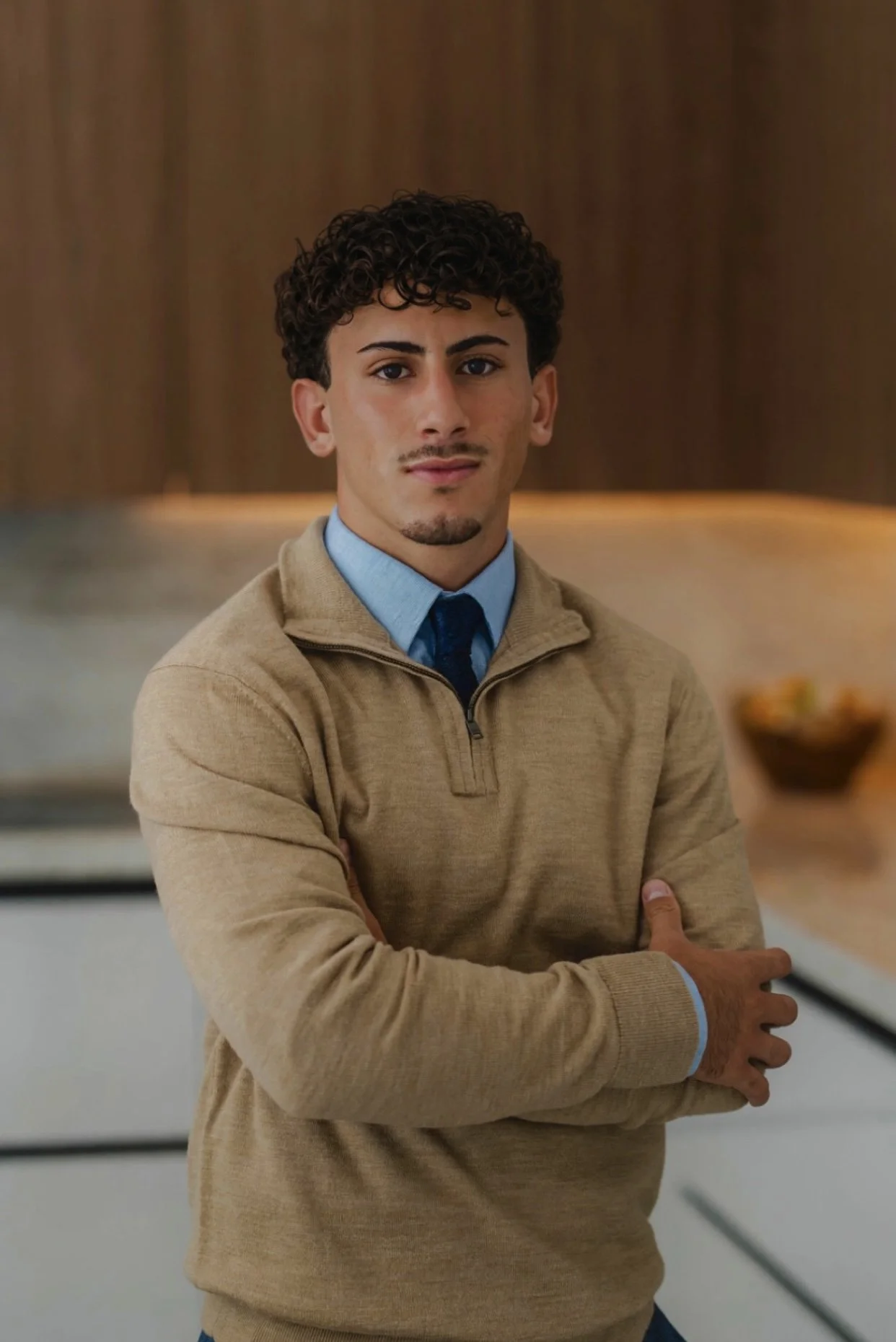 Young man with curly dark hair, wearing a beige sweater over a blue collared shirt and dark tie, standing with arms crossed in modern indoor setting.