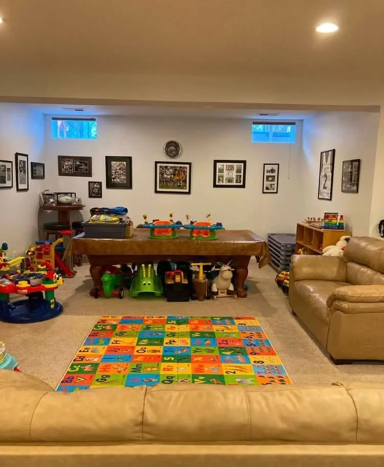 A basement playroom with a colorful alphabet rug, toys, and framed pictures on the wall. There is a leather couch and a pool table with children's toys underneath, and windows near the ceiling letting in natural light.