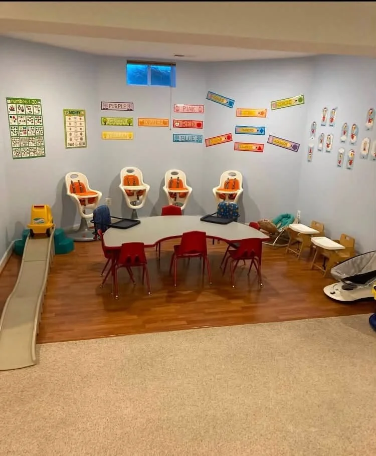 A preschool classroom corner with chairs, a table, a small slide, high chairs, and educational posters on the wall.