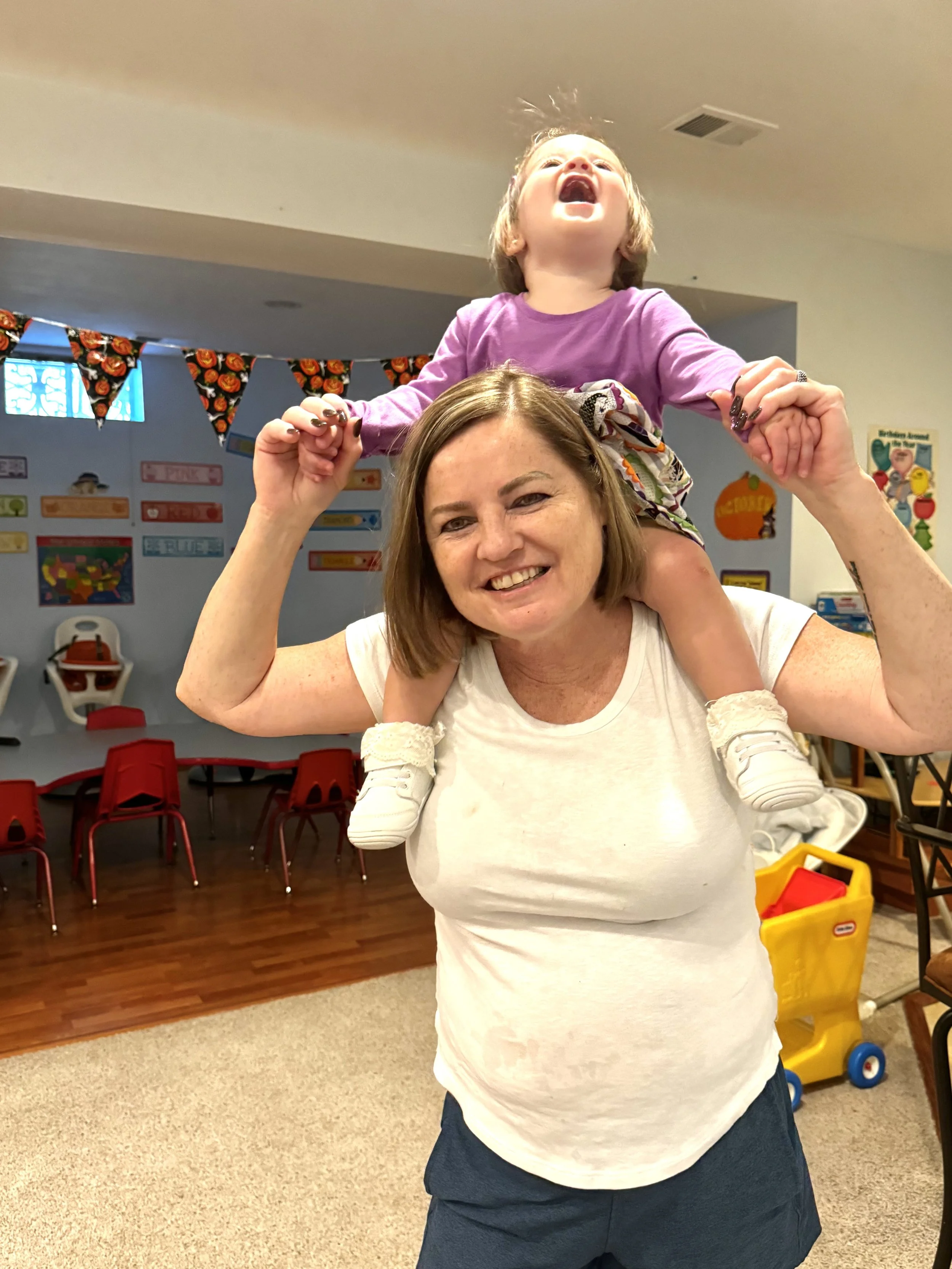 A woman smiling and holding a young girl on her shoulders, who is laughing with her mouth open. The background shows a room decorated for Halloween, with orange and black buntings and Halloween-themed posters. The girl is wearing a purple shirt and patterned shorts, and the woman is wearing a white shirt. There are red chairs and a yellow toy cart in the room.