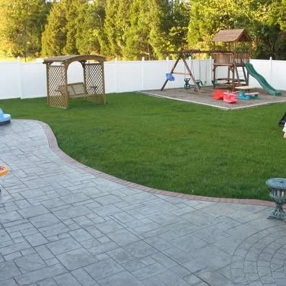 Backyard with green grass, a white fence, and a children's playset with a slide, swings, and a sandbox. There is a patio with a stone pattern.