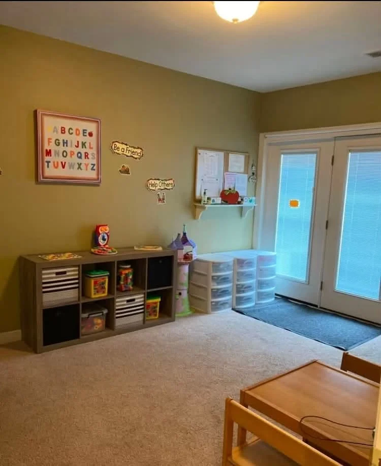 A children's playroom with a beige carpet, green walls, and sliding glass doors. The room has a wooden shelf with colorful toys, a white plastic storage unit, and wall decorations including an alphabet chart and motivational signs. There's also a small wooden table with chairs.