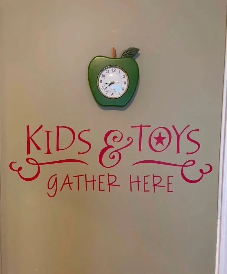 A wall with a green apple-shaped clock at the top and red and pink text that reads 'Kids & Toys Gather Here'.