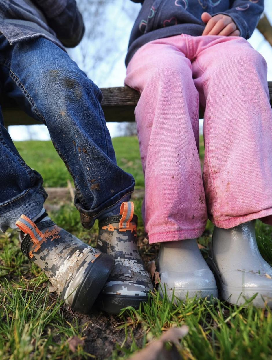 Close-up of two children sitting outdoors on a wooden bench, wearing muddy boots and colorful pants, in a grassy area with trees in the background.