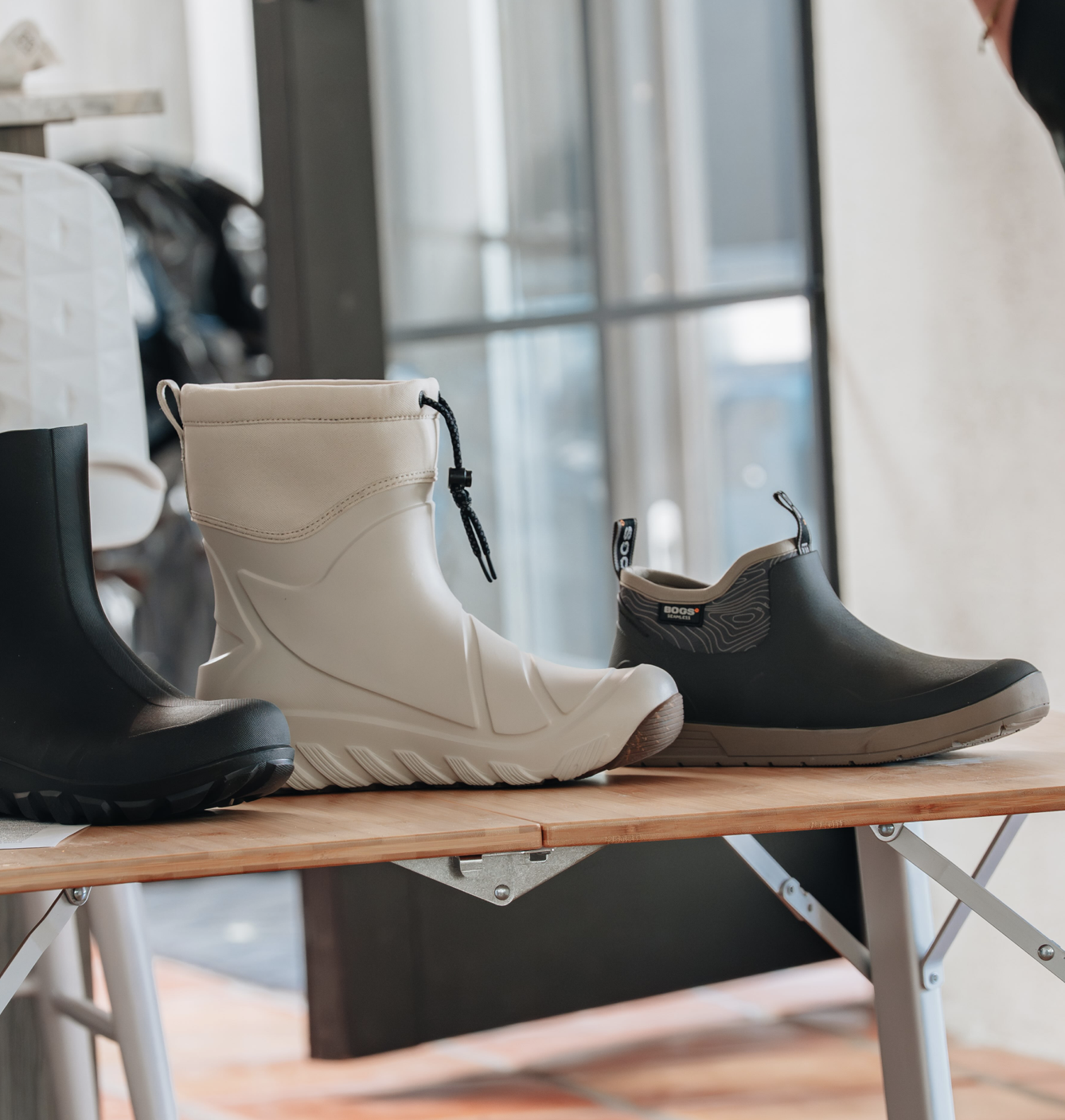Three pairs of rubber boots on a wooden table, with a black, white, and gray pair, in a bright room with large windows.